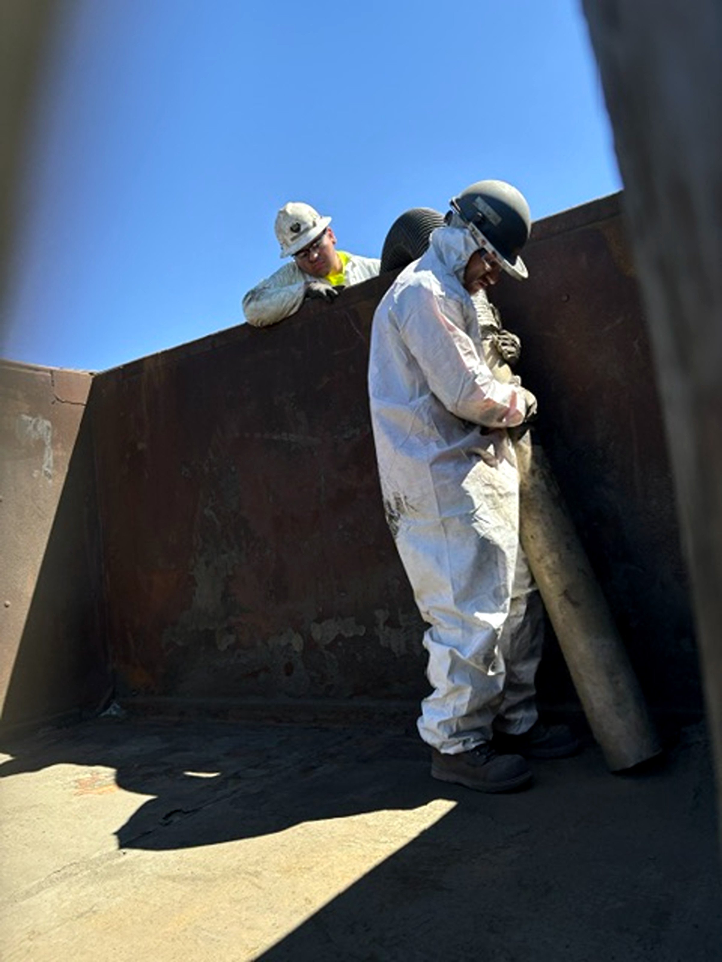 Two Prime Industrial workers in Northern Nevada wearing helmets and safety gear pressure washing inside a large, metal mine equipment structure.