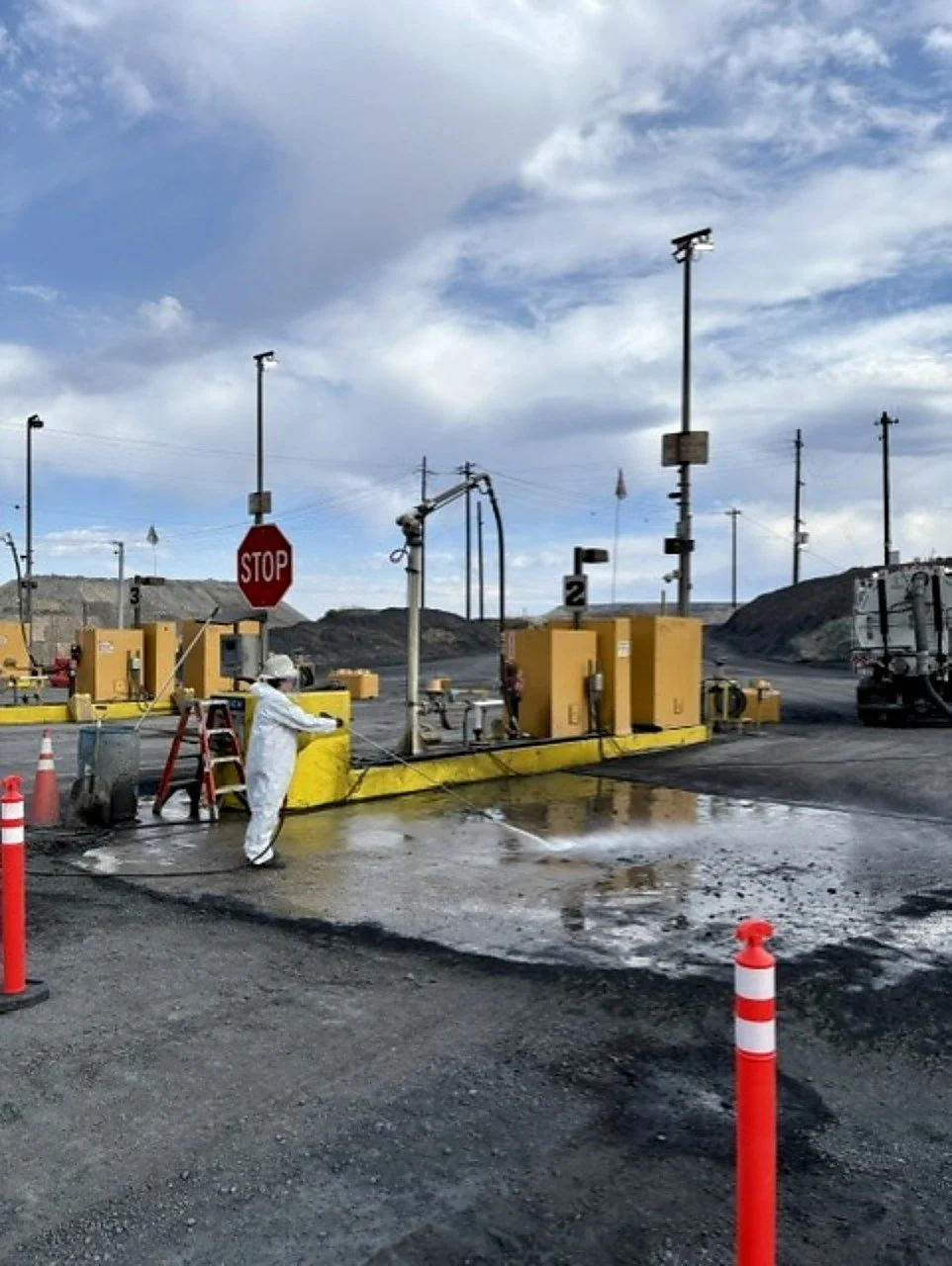 Prime Industrial employee in Northern Nevada wearing a white protective suit and helmet cleaning a fueling station or industrial site with water, amid gravel and dirt ground, outdoor setting under cloudy sky.