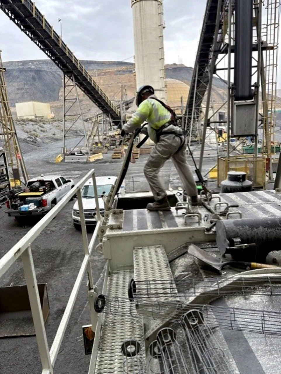 Construction worker wearing safety gear, including a helmet and reflective vest, standing on a metal platform at an industrial site with large machinery, conveyor belts, and trucks around.