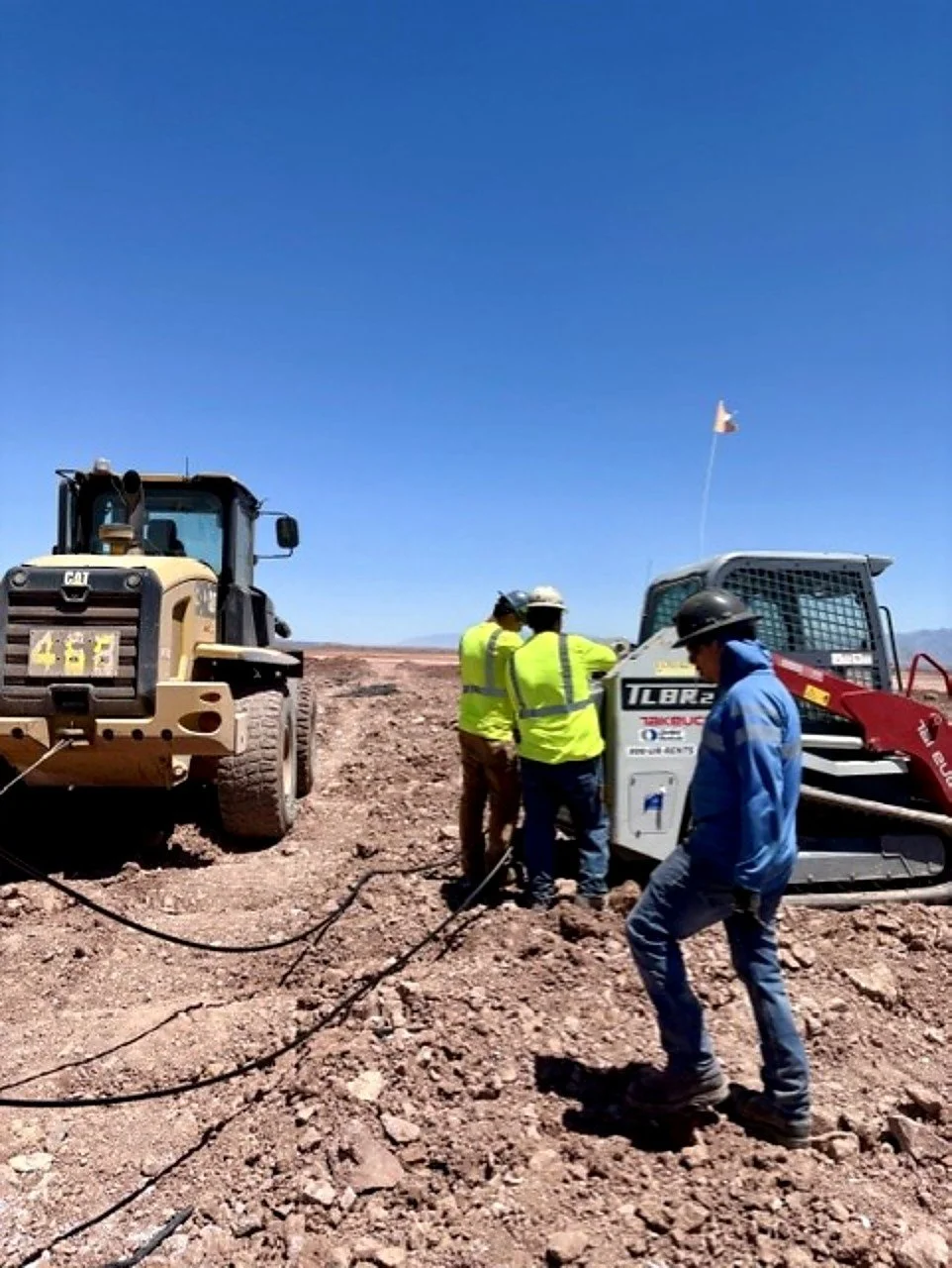 Prime Industrial employees in Northern Nevada wearing yellow and blue safety gear work around a small skid-steer loader in a rocky, open landscape under a clear blue sky.