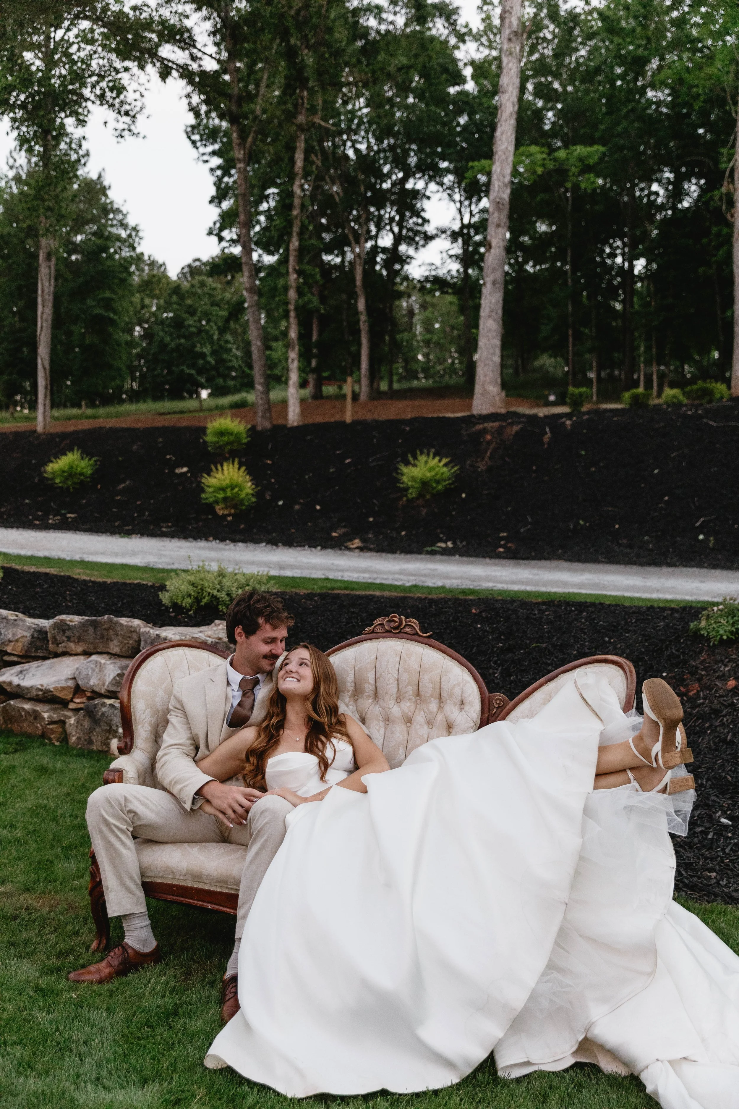 A couple dressed in wedding attire sitting on a vintage sofa outdoors, with the bride resting her head on the groom's shoulder, smiling and looking at each other, surrounded by a lush green landscape.