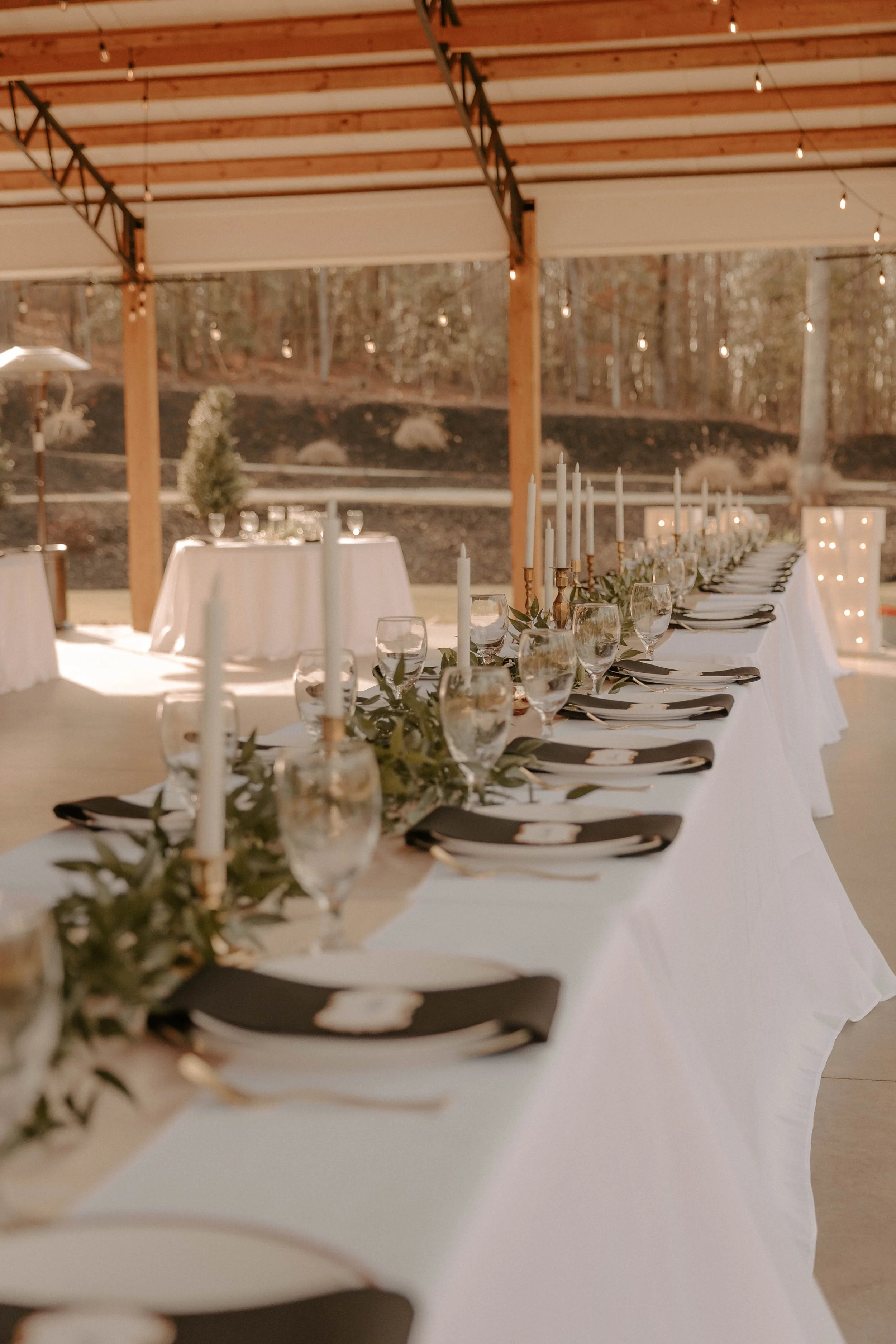 A long banquet table covered with a white tablecloth, set with wine glasses, plates, and silverware, decorated with a green leafy garland, candles, and surrounded by wooden beams with hanging string lights. An outdoor setting with trees and a hillside in the background.