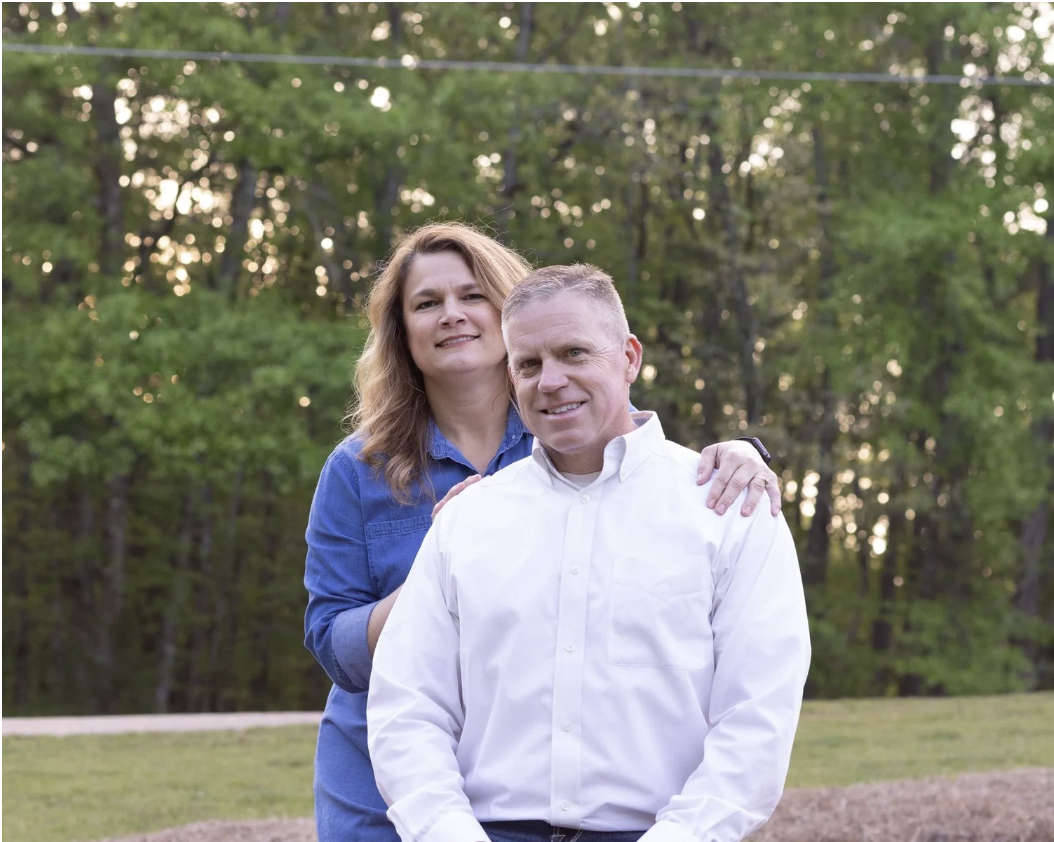 A man and woman outdoors in a park with trees in the background. The woman stands behind the man, resting her hands on his shoulders, both smiling at the camera.