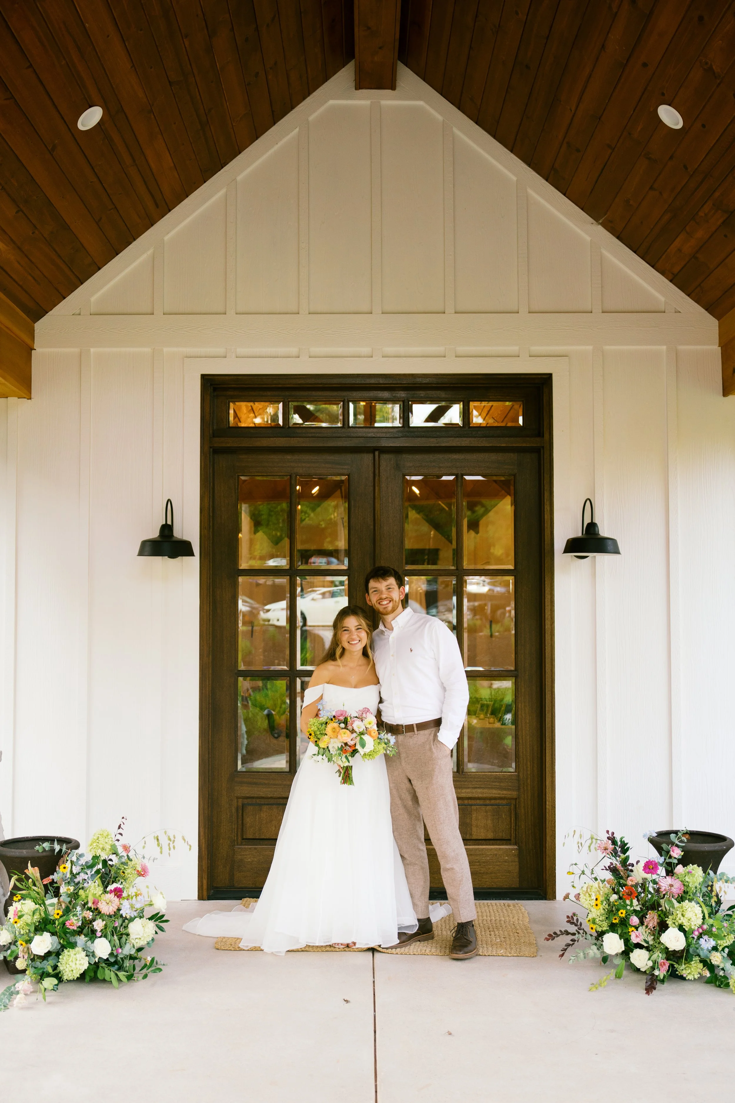 A smiling bride and groom standing together outside a house with a dark wood door, surrounded by colorful flower arrangements on the ground.
