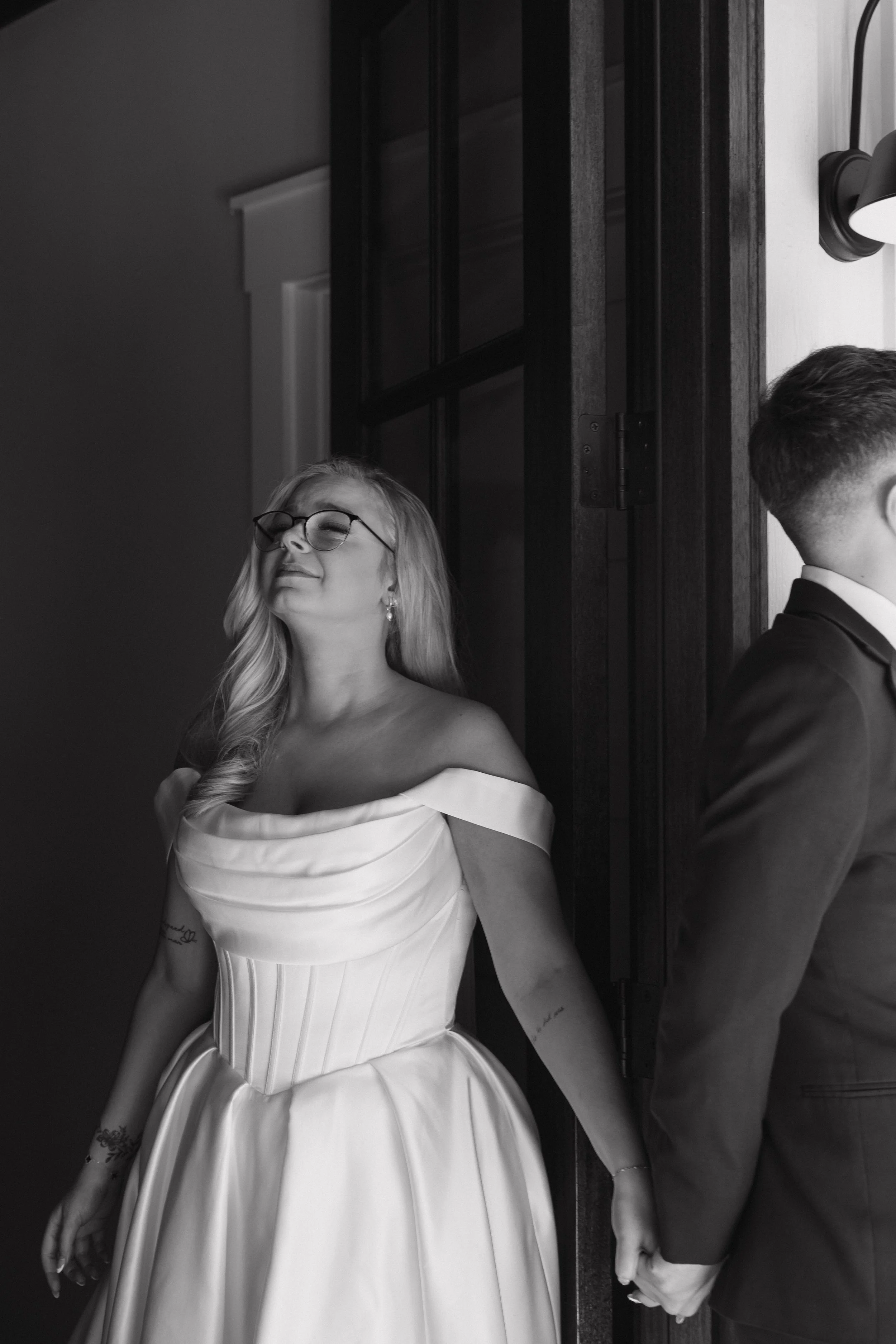A woman in an elegant off-the-shoulder white dress with a fitted bodice and flowing skirt, smiling with her eyes closed, and wearing glasses, holding hands with a man in a suit, standing beside her in front of a dark wooden door.