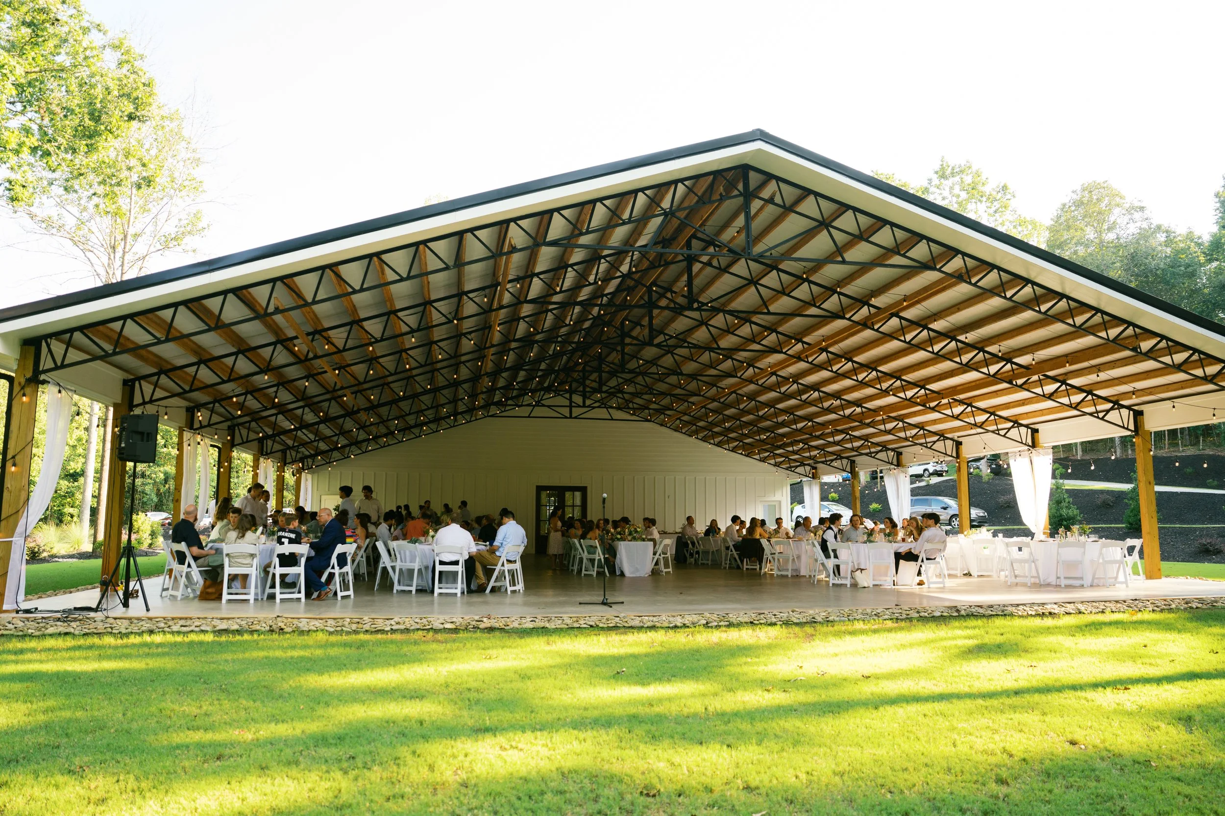 Outdoor wedding reception under a large open pavilion with string lights and surrounded by trees, with guests seated at round tables.