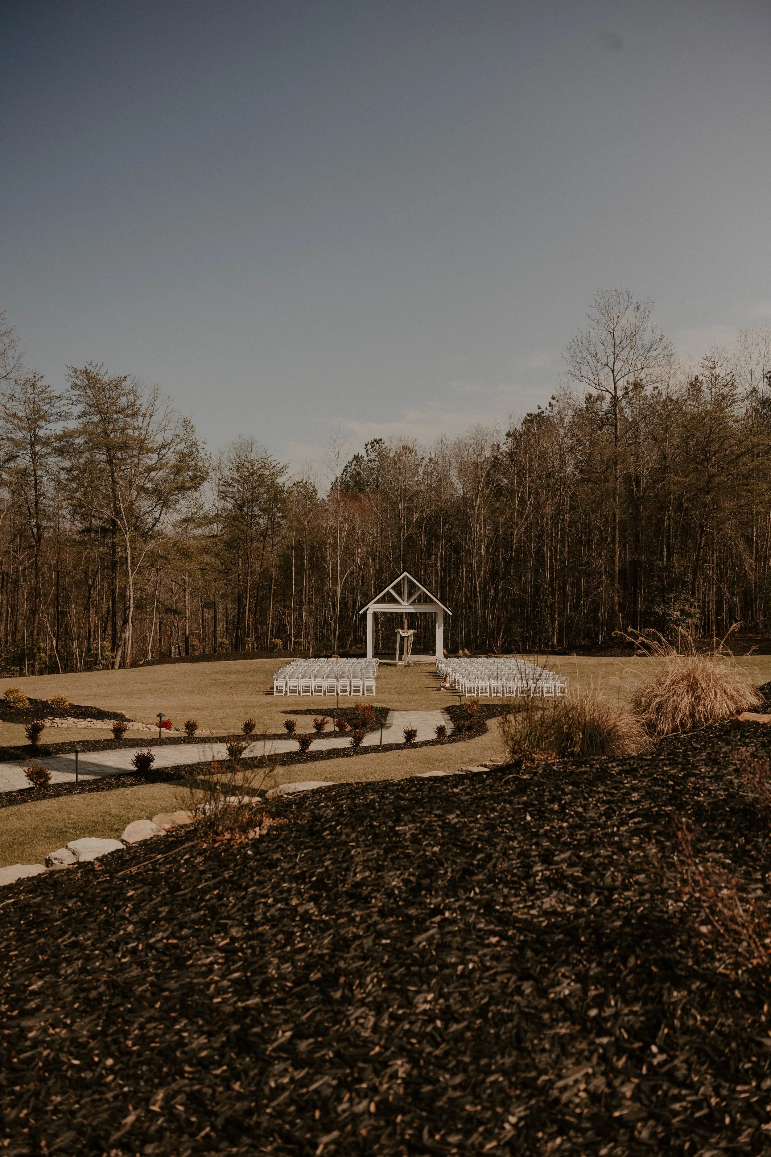 A small white open-air pavilion with a cross and a hanging wreath, set on a grassy area surrounded by white chairs, against a background of leafless trees under a cloudy sky.