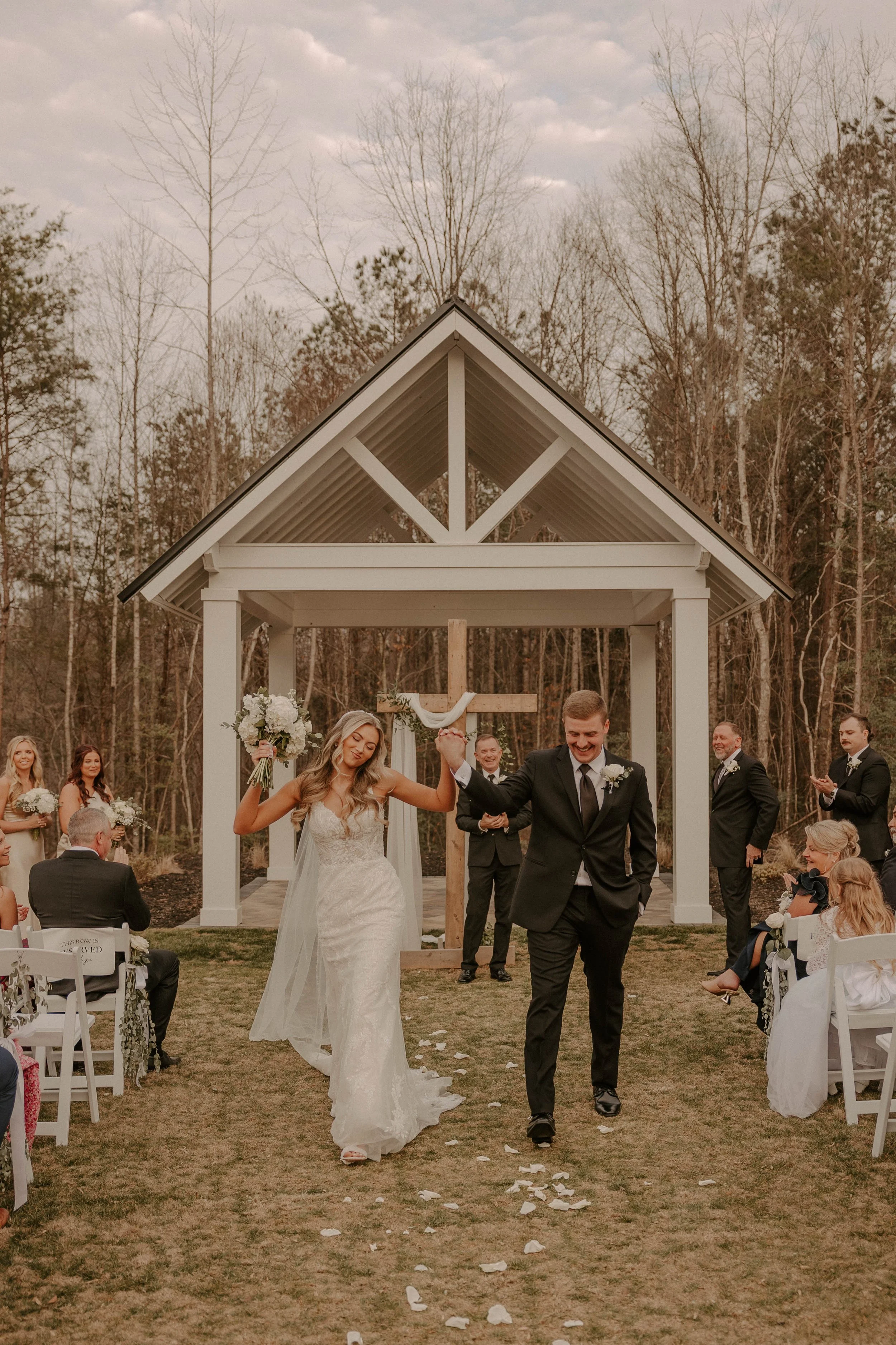 A bride and groom walk down the aisle at their outdoor wedding ceremony, surrounded by friends and family with trees in the background.