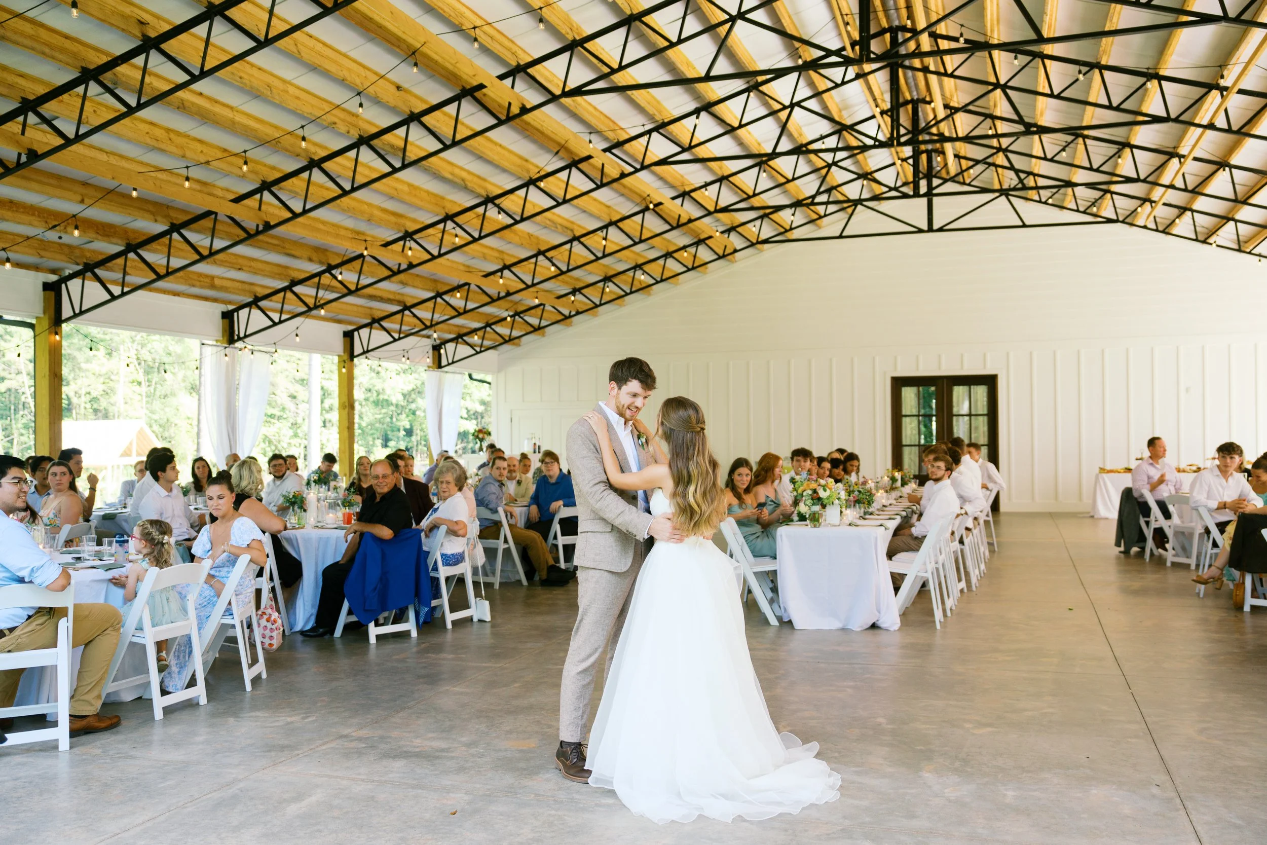 A bride and groom are dancing at their wedding reception in a spacious hall with a high wooden ceiling and string lights. Guests are seated at tables decorated with flowers, watching them dance.