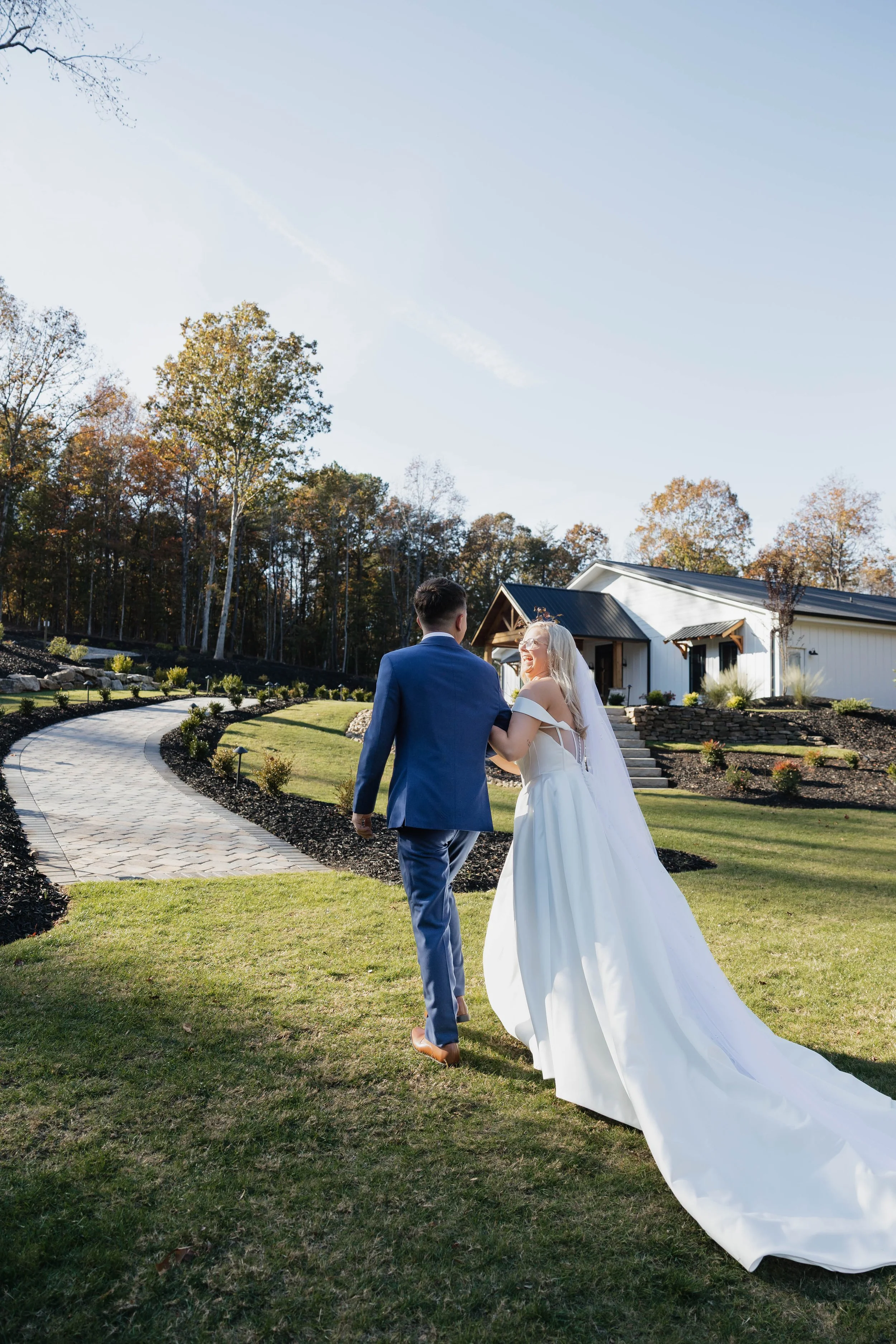 A bride and groom walking hand in hand on a stone pathway outside a white house with a black roof, surrounded by trees with autumn foliage.