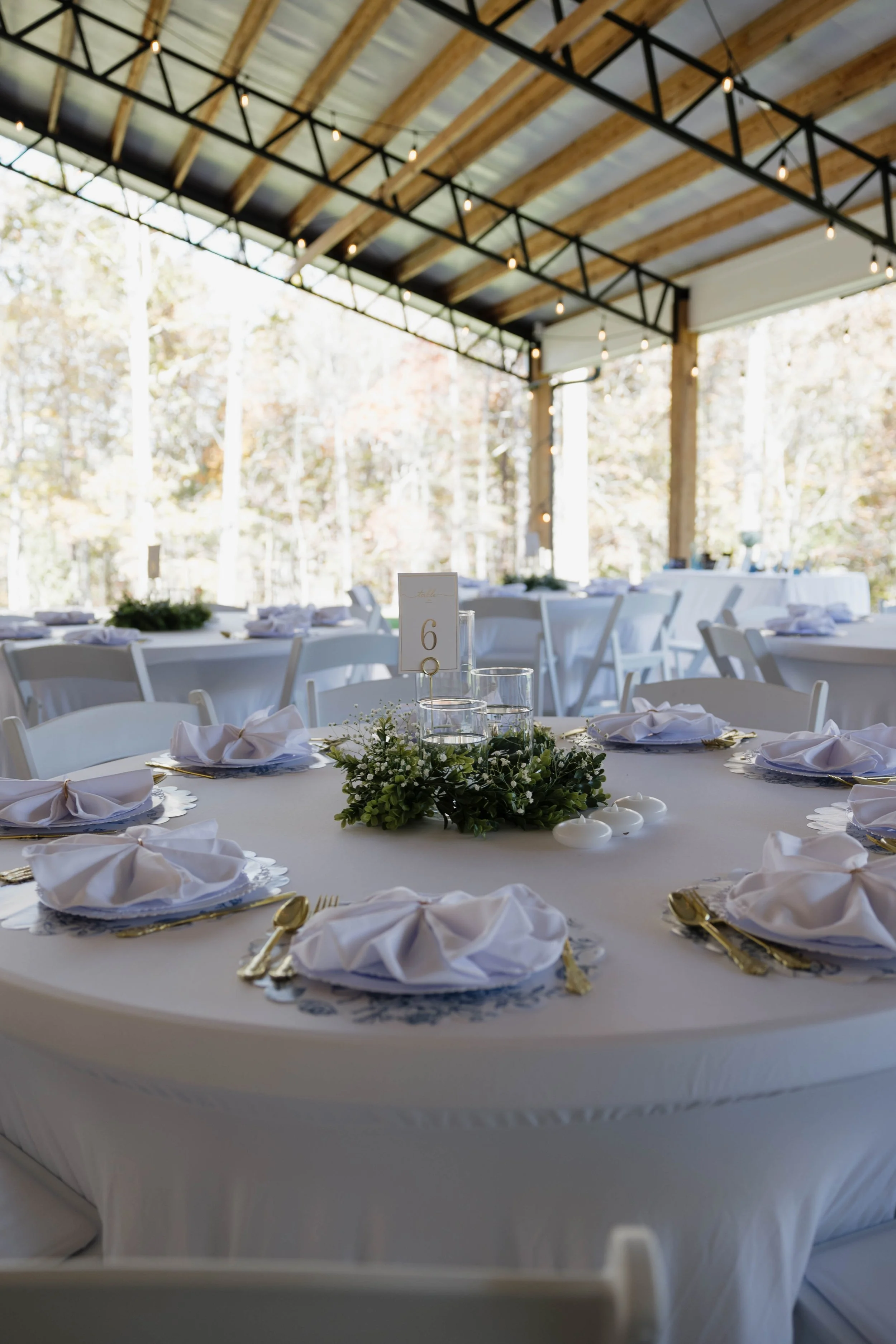 Indoor wedding reception with round tables covered in white tablecloths, gold utensils, white napkins folded on plates, and floral centerpieces. The ceiling has exposed wooden beams and string lights, with large windows showing trees outside.