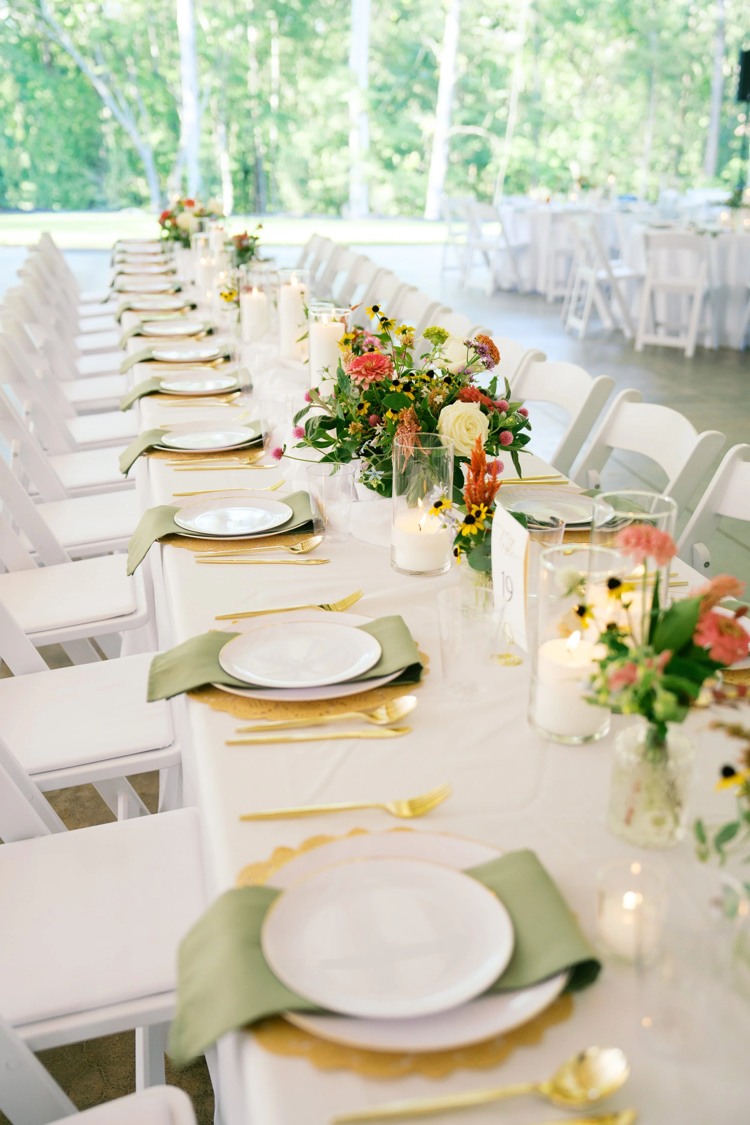A long banquet table decorated for a celebration, with white chairs, floral centerpieces, candles, and tableware, set in a bright, outdoor setting surrounded by trees.