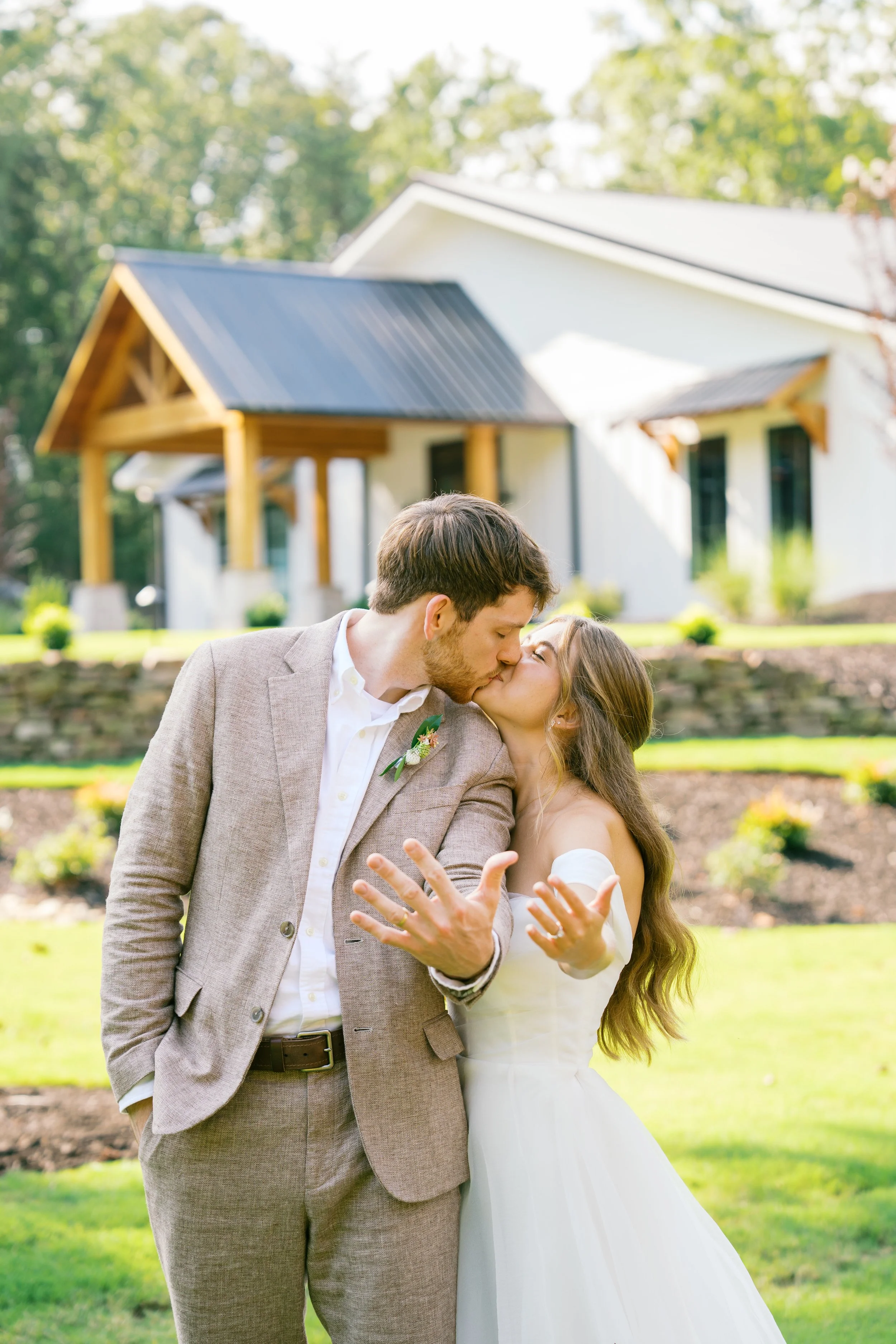 A wedding couple, a man and woman, sharing a kiss outdoors in front of a white house with a porch, surrounded by green grass and trees.