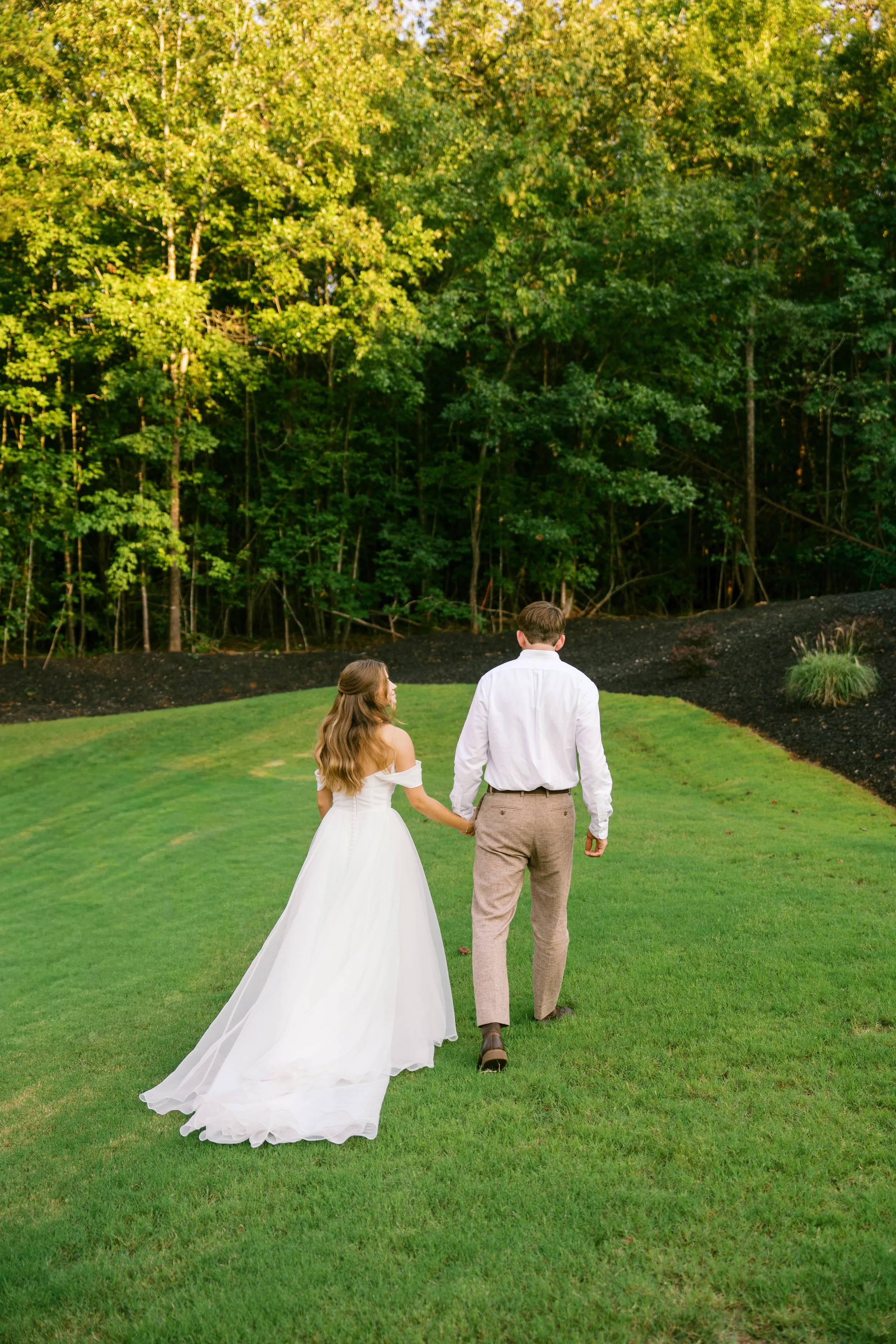 A couple dressed in wedding attire walking away from the camera on a grassy lawn with a dense forest in the background.