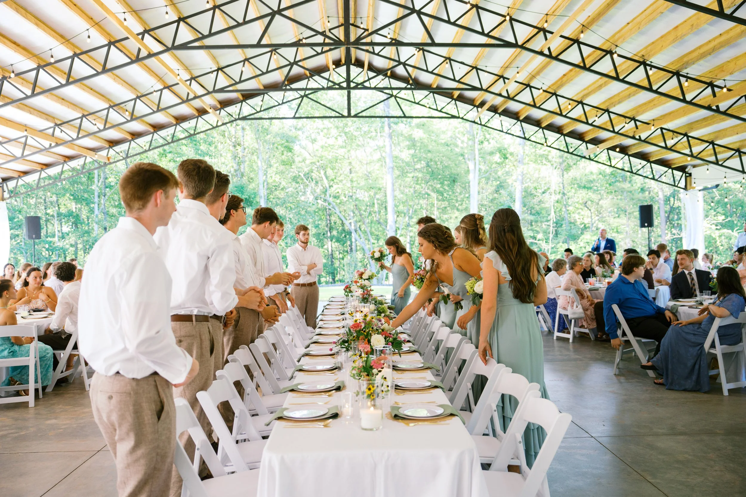 People gathered at a long dining table during a wedding reception or celebration under a covered outdoor pavilion with string lights, surrounded by trees.