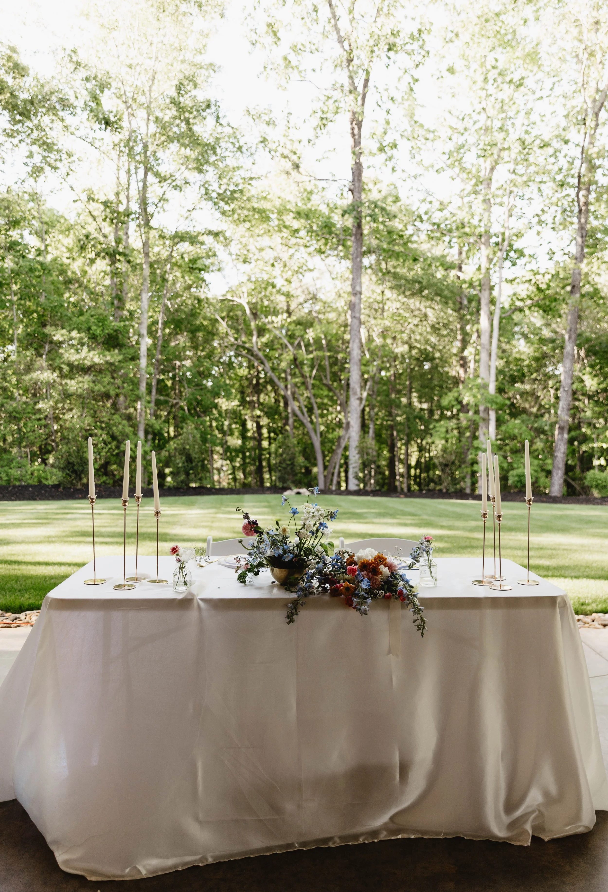 A long rectangular table with a white tablecloth, decorated with flower arrangements and tall white candles, set outdoors in a green wooded area.