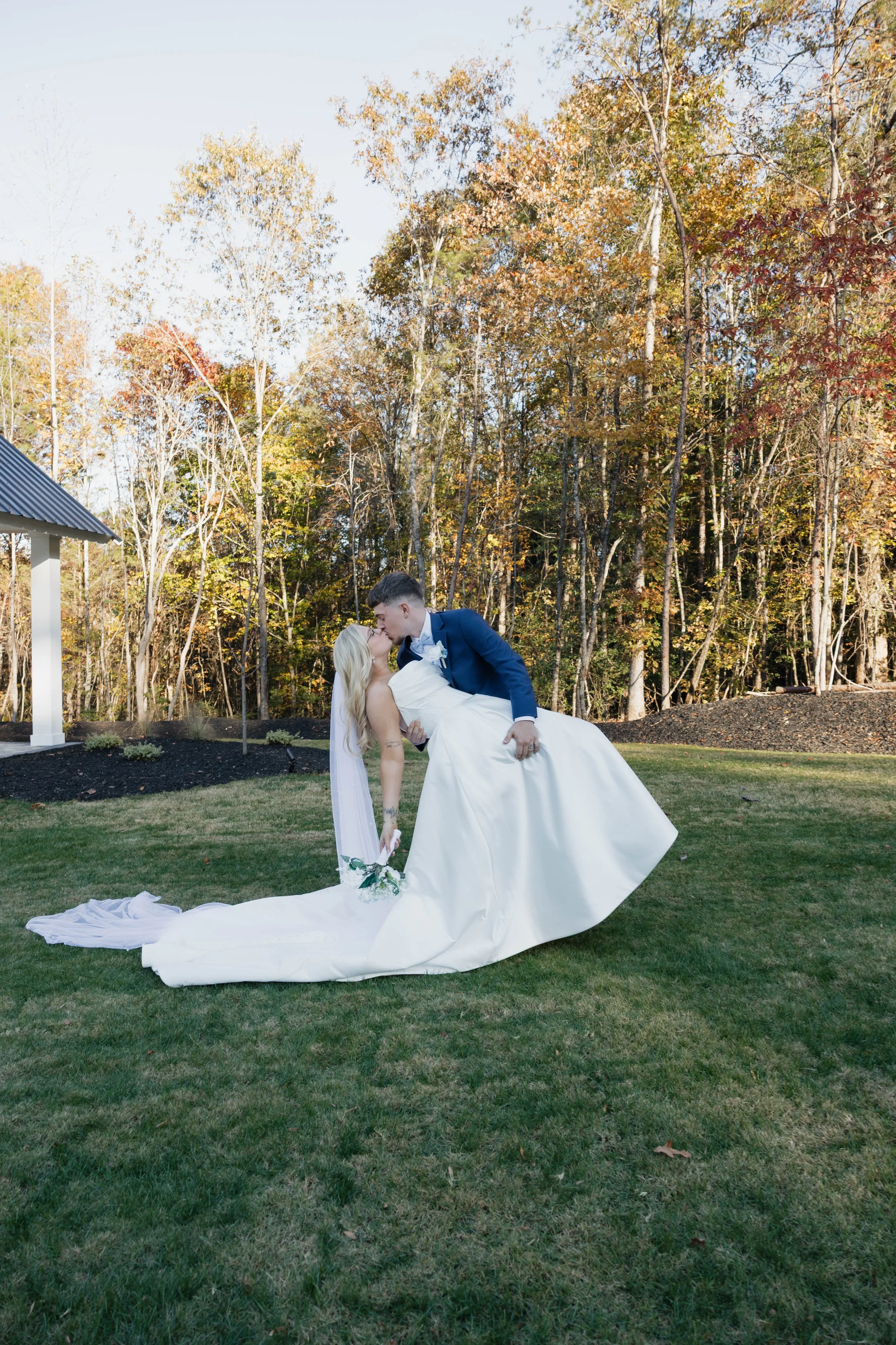 A newlywed couple sharing a kiss outdoors with a backdrop of trees with fall foliage. The groom is dipping the bride, who is holding a bouquet, on a grassy lawn.
