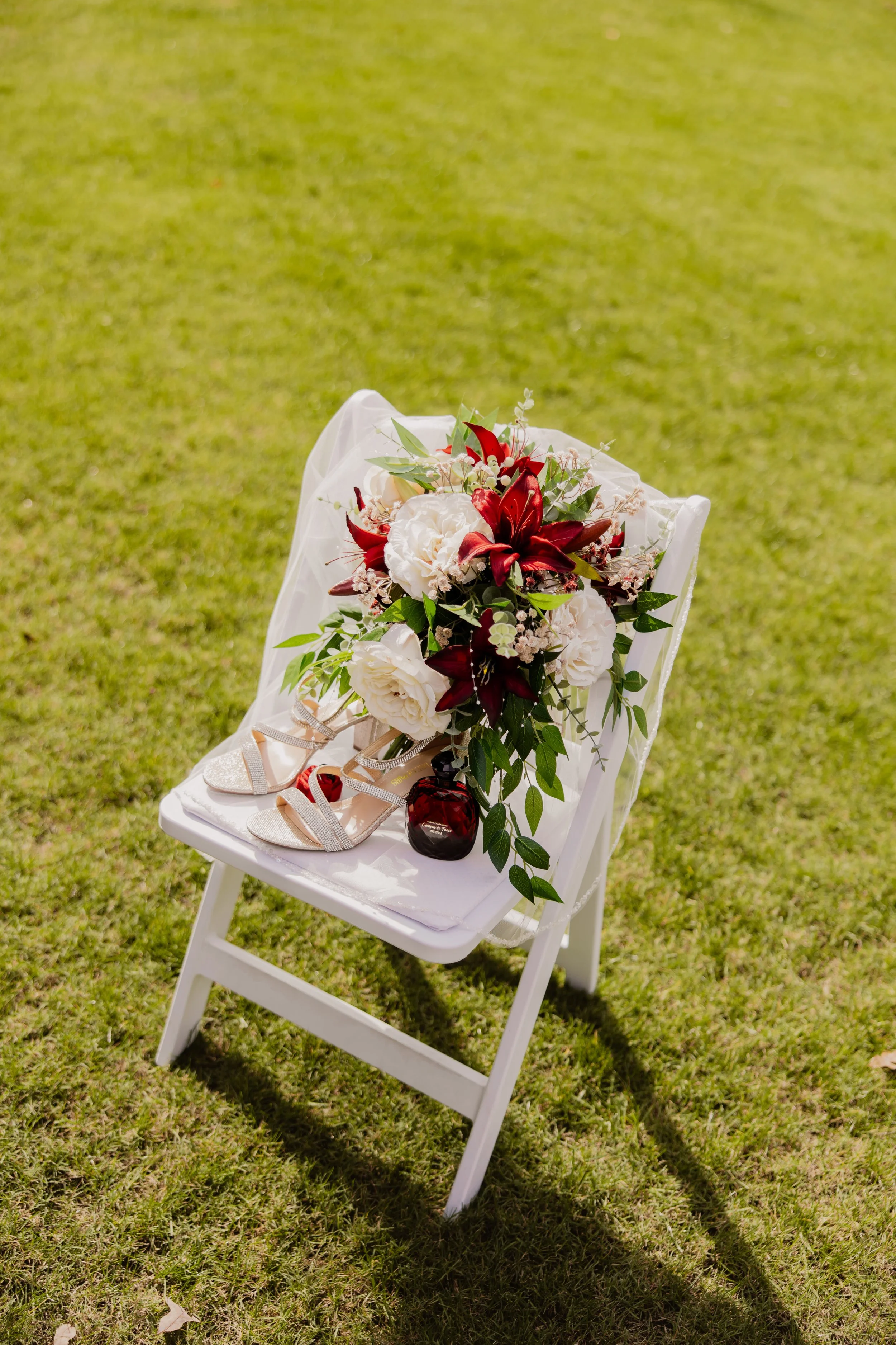 A white folding chair on green grass decorated with a floral arrangement of white and red flowers and greenery, with a pair of glittery shoes and a small bottle placed on the chair's seat.