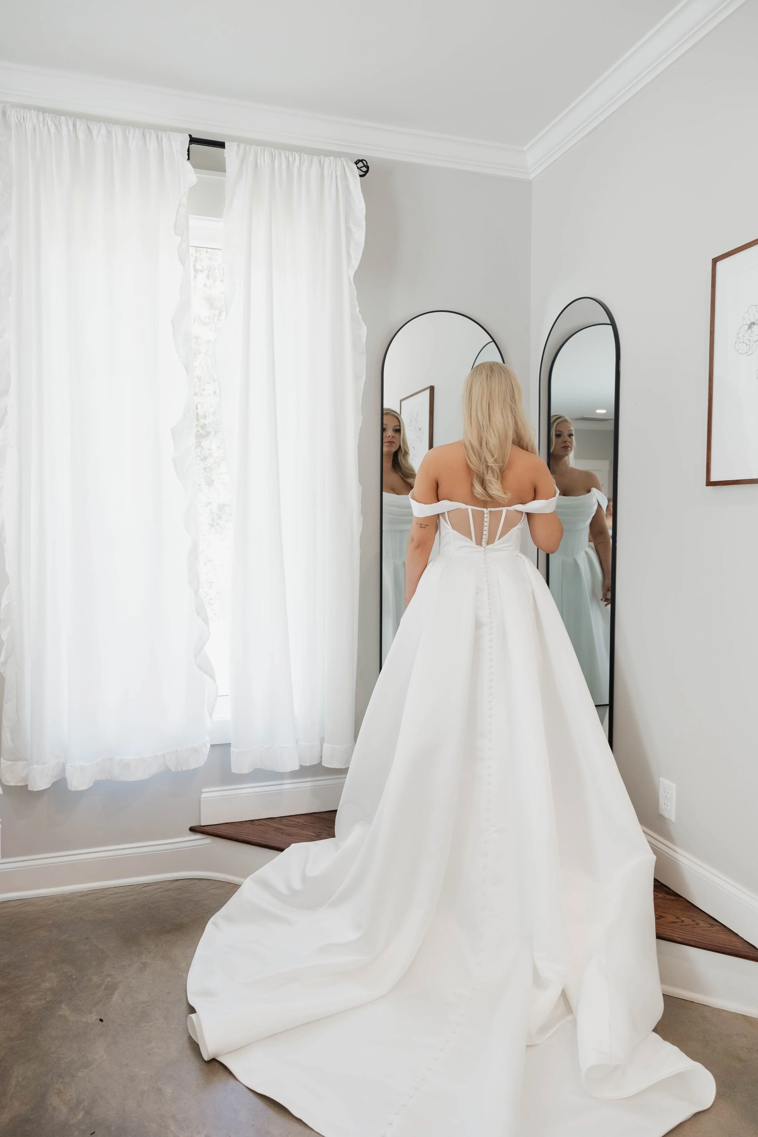 A woman in a white wedding dress stands in front of two tall mirrors, looking at her reflection. The room features white walls, white curtains, and framed artwork. The wedding dress has an off-the-shoulder design with lace-up back detail, and the tra