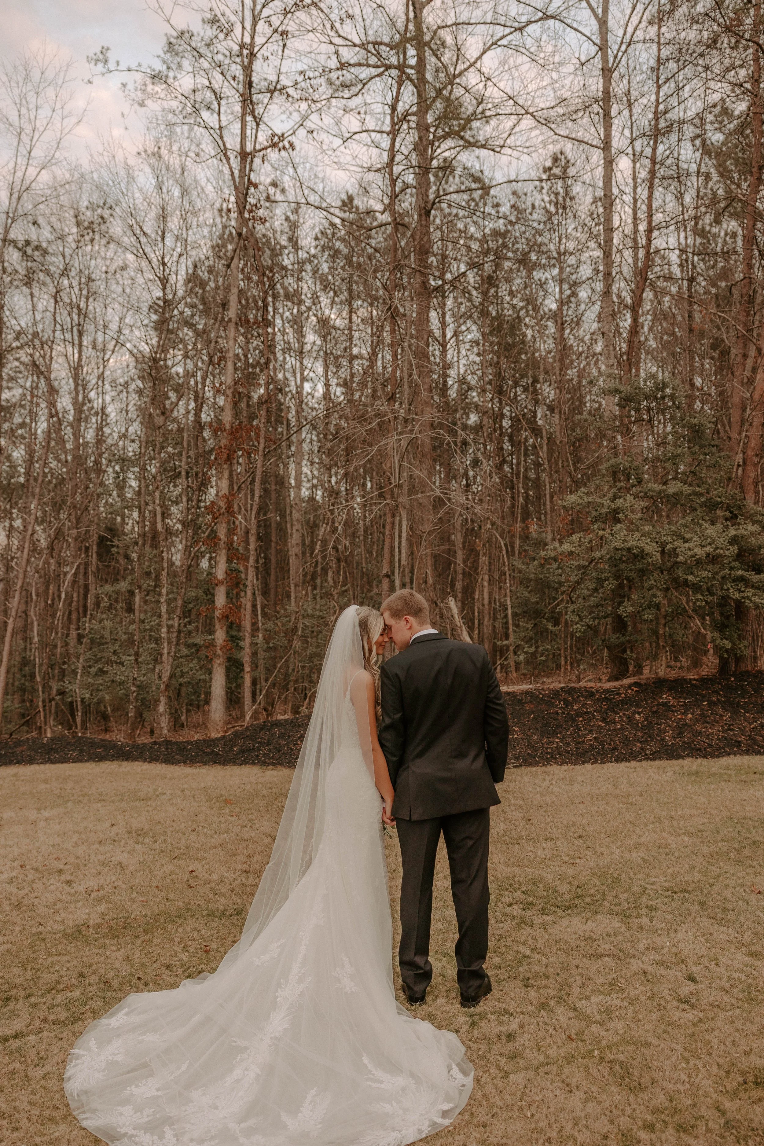 A bride and groom holding hands with foreheads touching, standing outdoors on a grass lawn with a forest in the background, during a wedding ceremony.