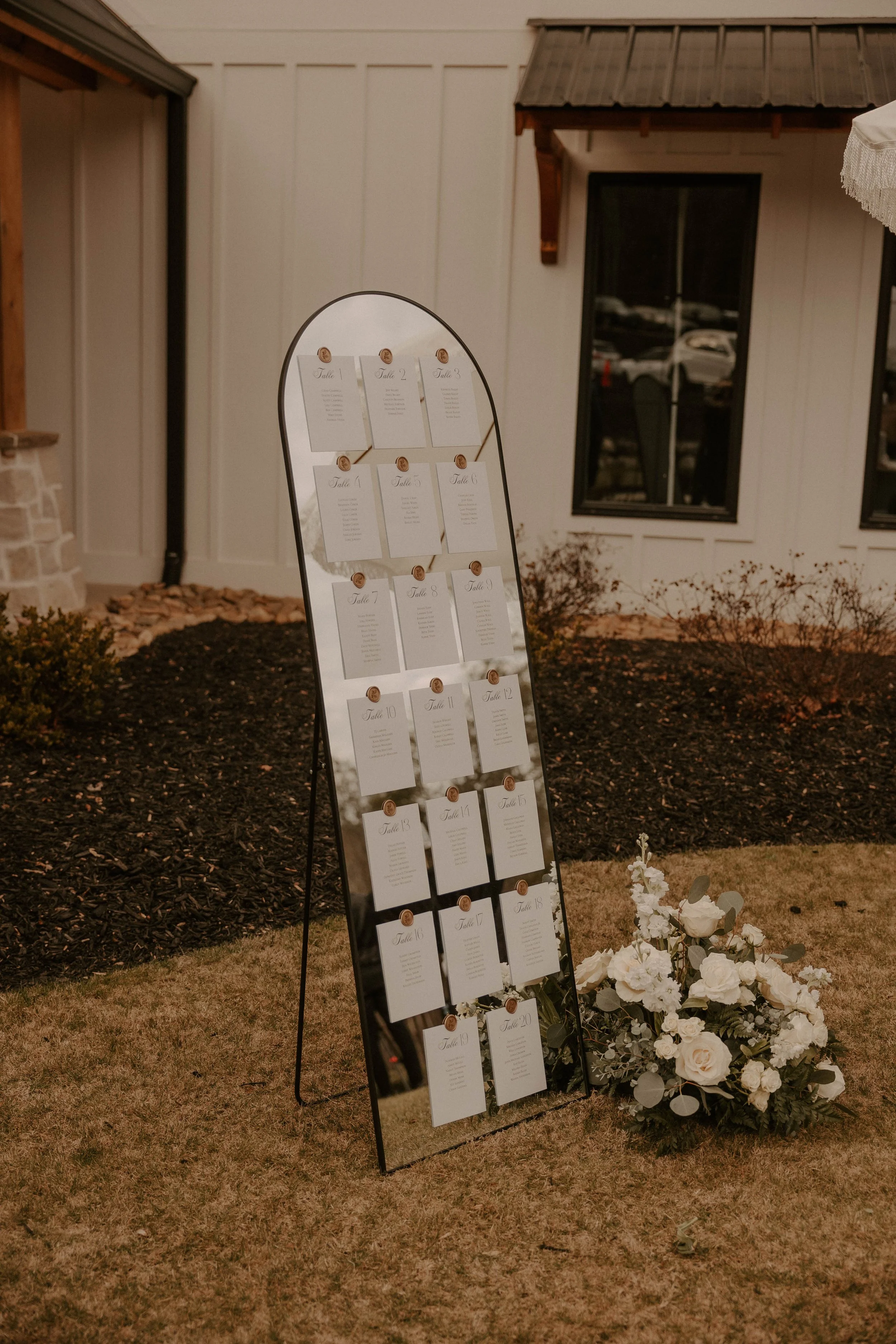 A large, curved mirror-style wedding seating chart with 20 labeled sections from Table 1 to Table 20, set outdoors near a building with white siding and windows, decorated with white roses and greenery.