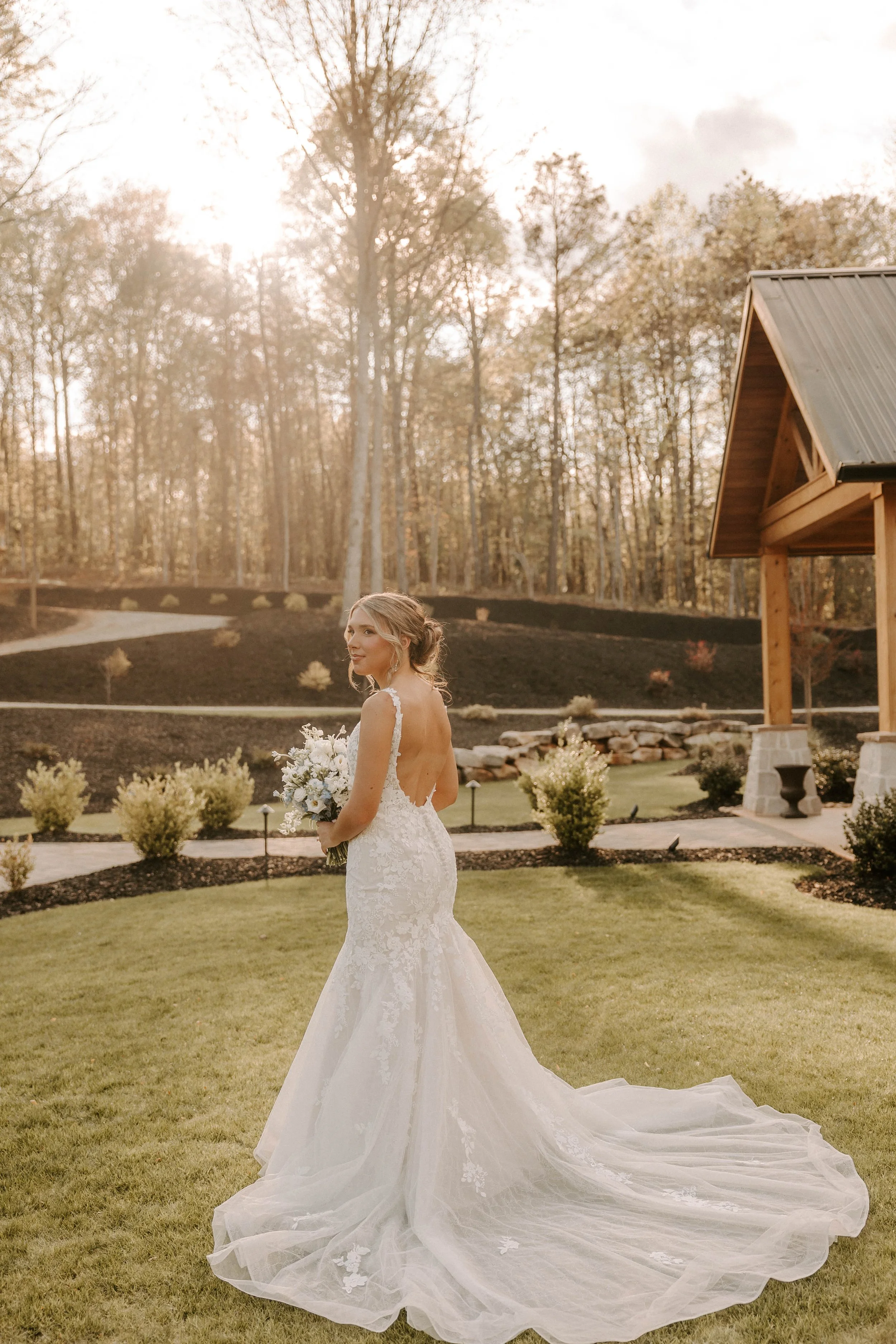 A bride in a white lace wedding dress holding a bouquet. She is outdoors during sunset with trees and a small wooden building in the background.