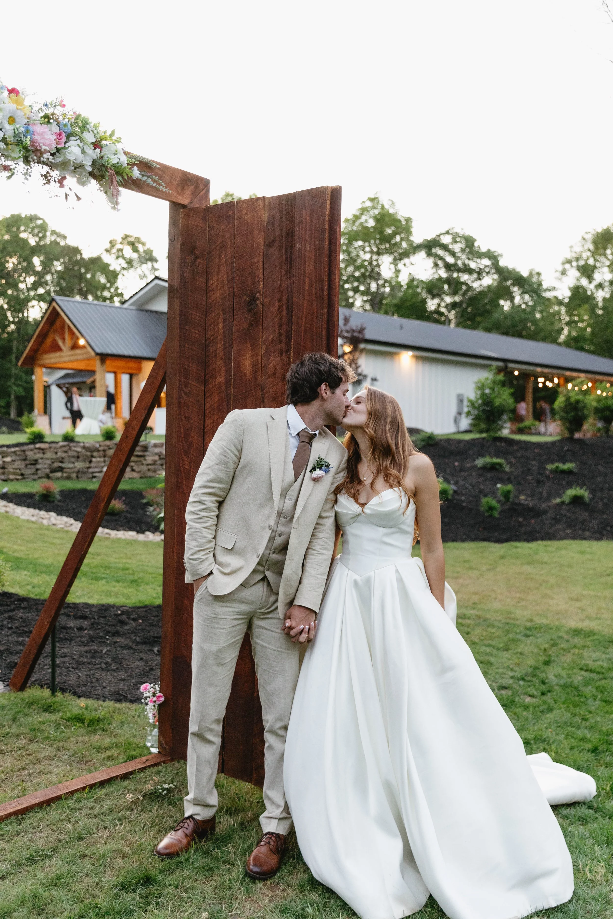 A couple in wedding attire kissing under a wooden arch with flowers, outdoors during the evening, with a building with string lights in the background.
