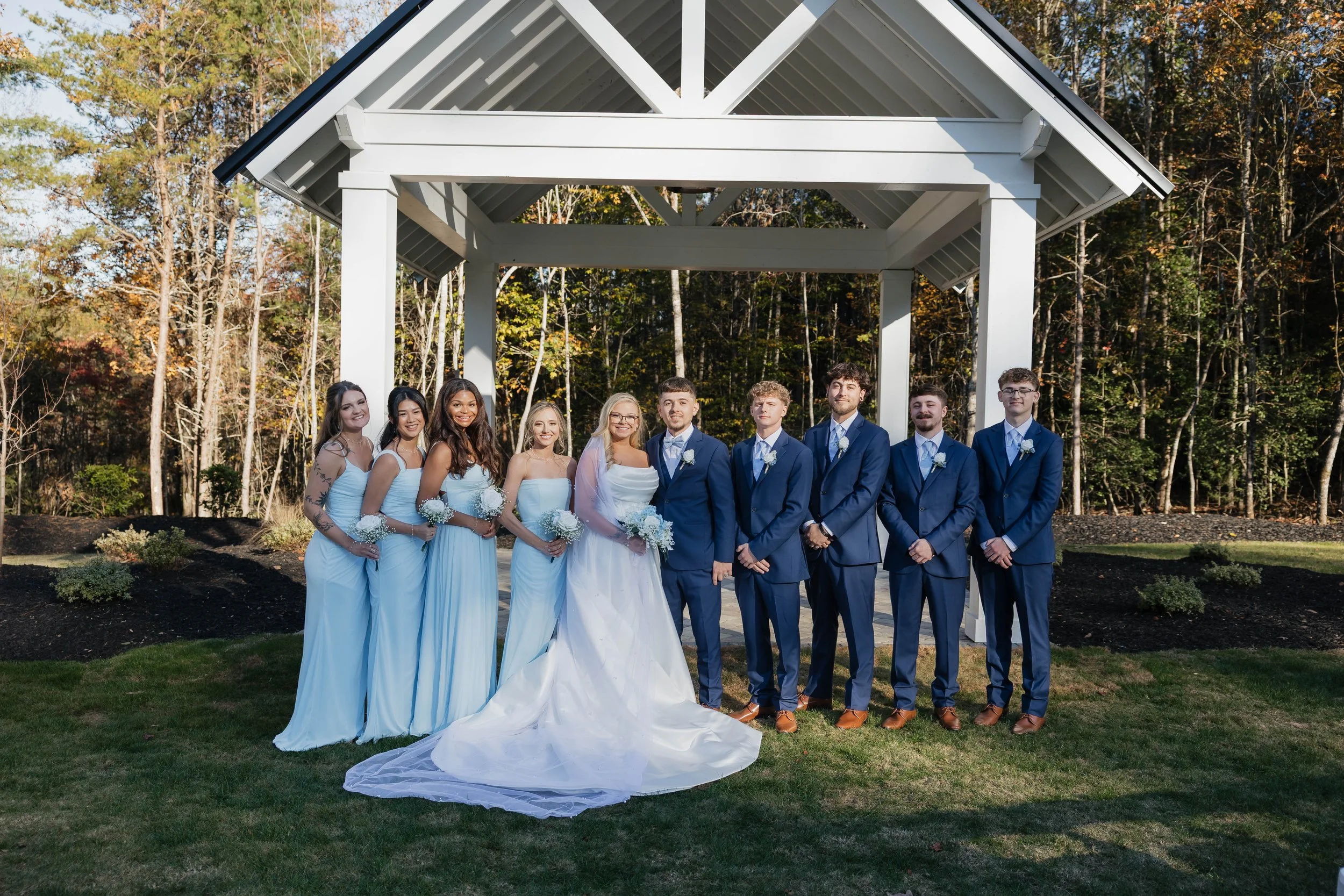 Group of wedding couples under a white gazebo in an outdoor setting with trees in the background.
