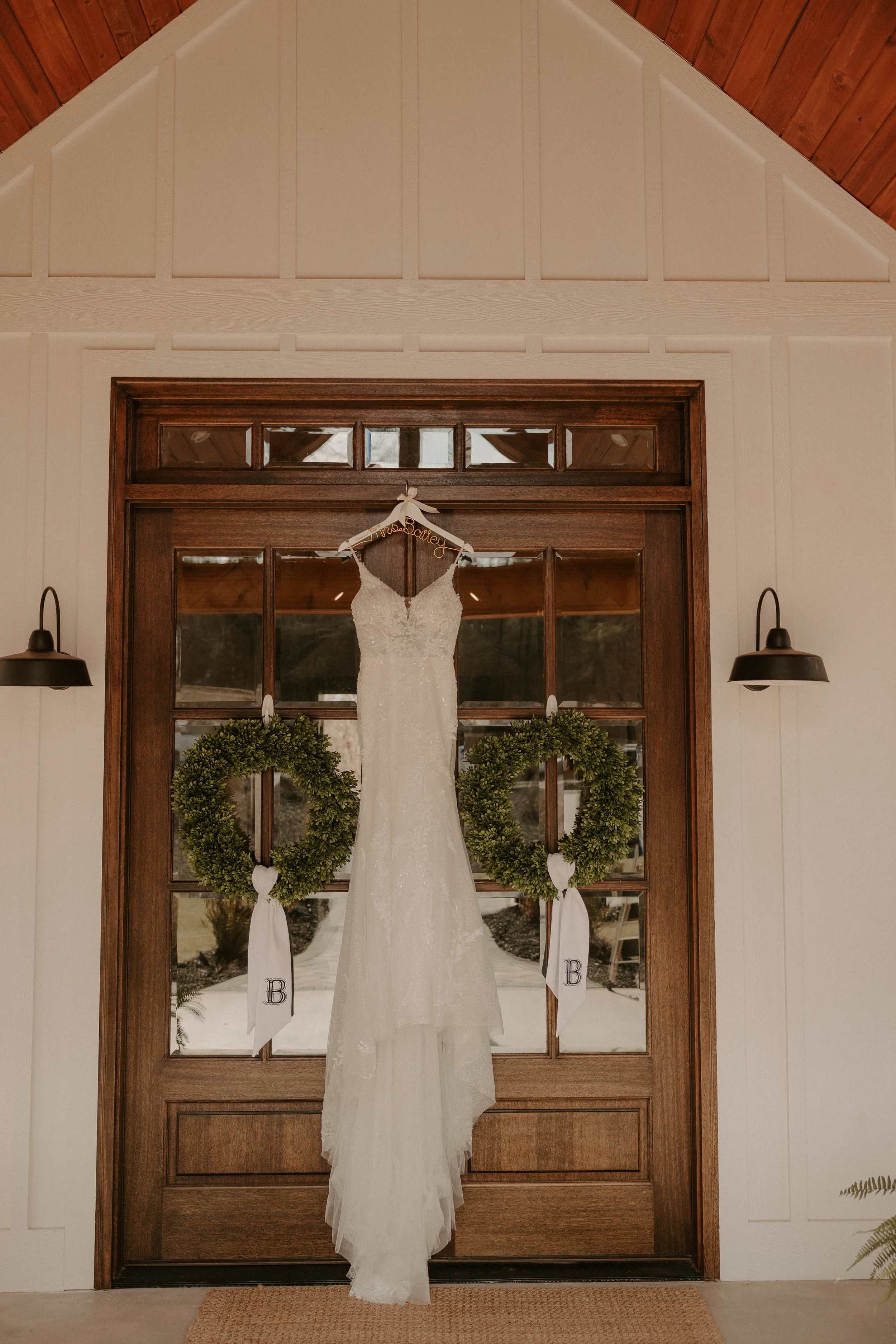 A wedding dress hanging on a hanger in front of a wooden door with glass panes, decorated with two green wreaths with white bows and lettered tags.