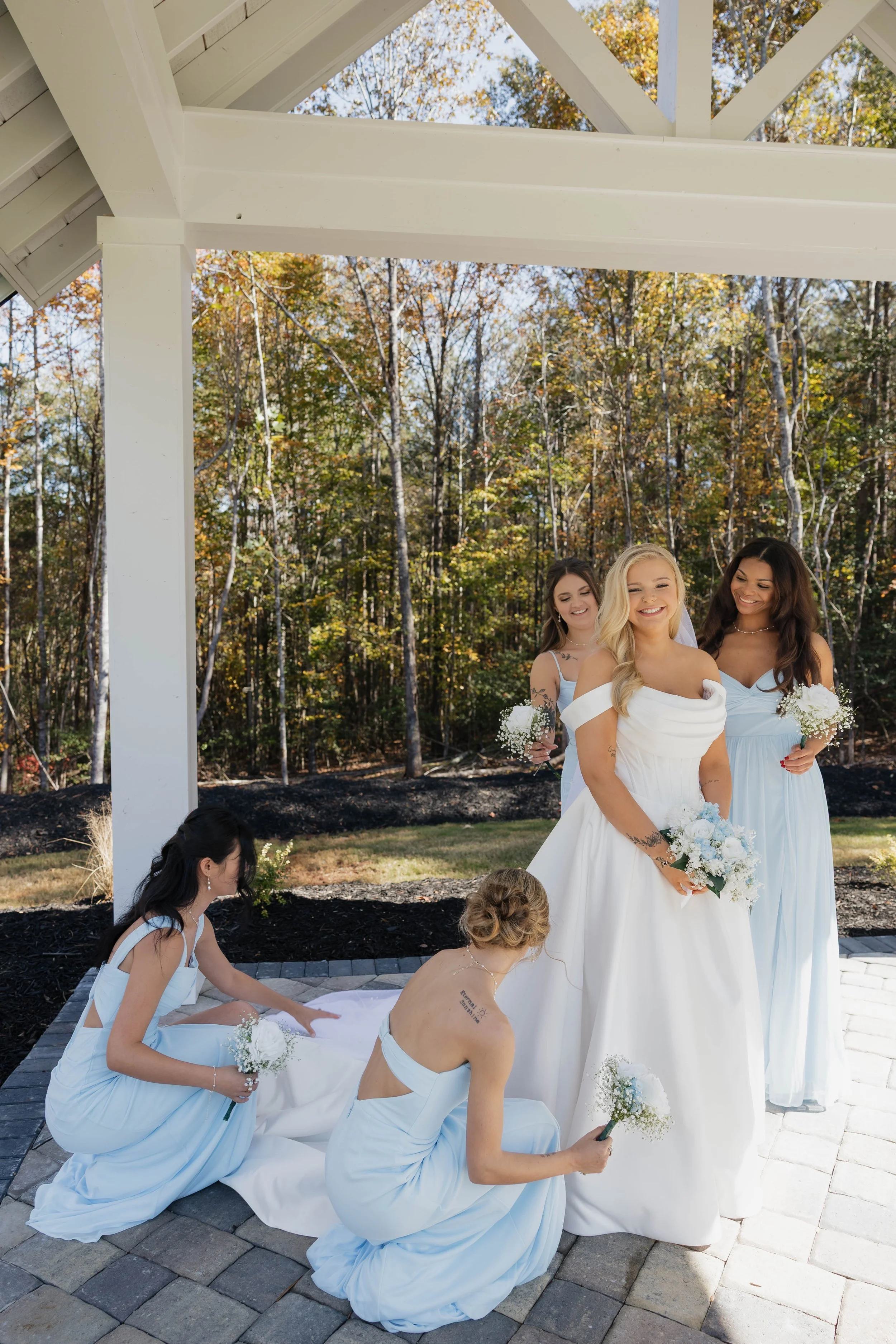 A group of women in light blue dresses and a bride in a white wedding gown getting ready for a wedding ceremony outdoors under a white wooden structure with trees in the background