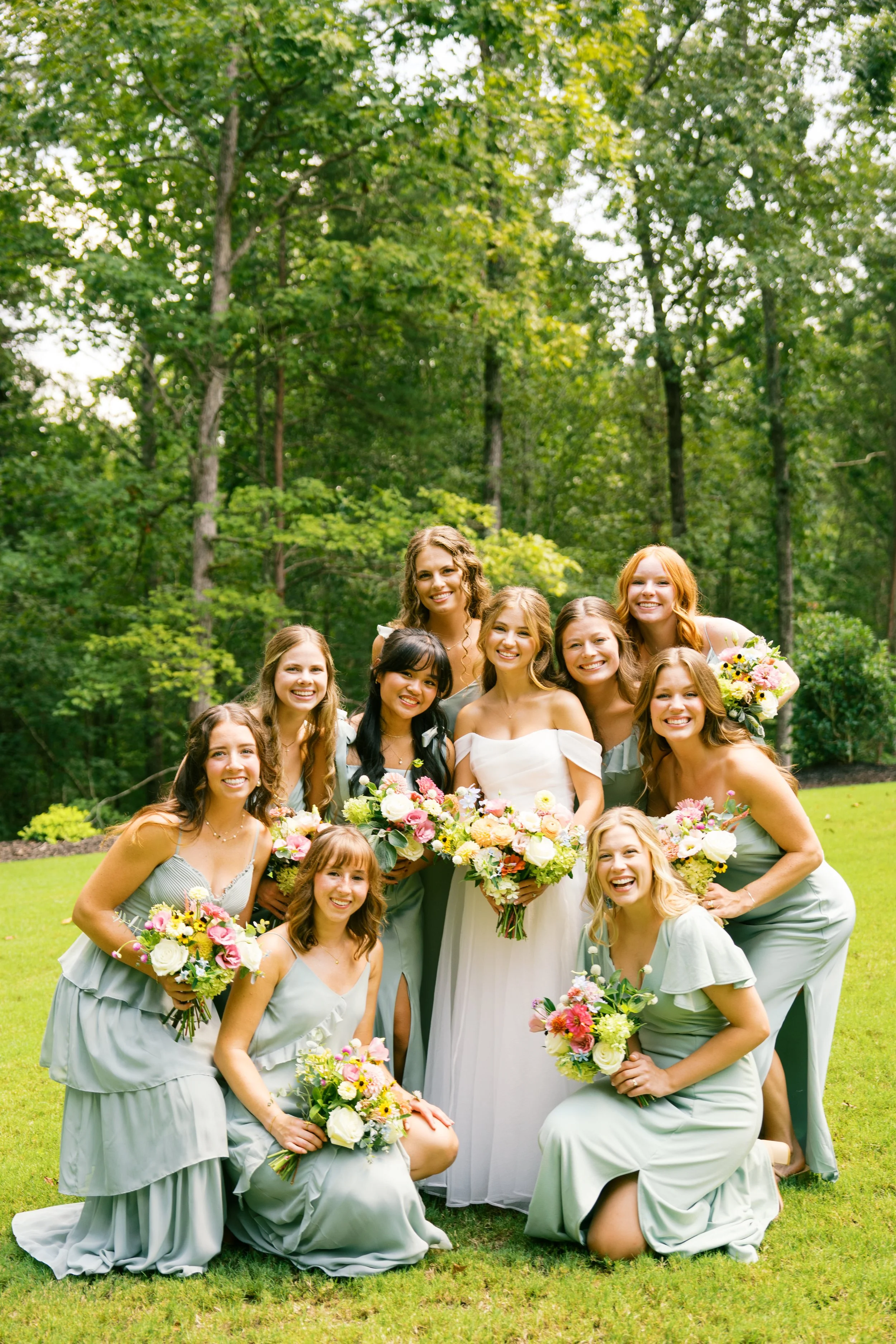 Group of women wearing matching grey dresses, posing outdoors with greenery in the background, holding colorful bouquets, celebrating a wedding.