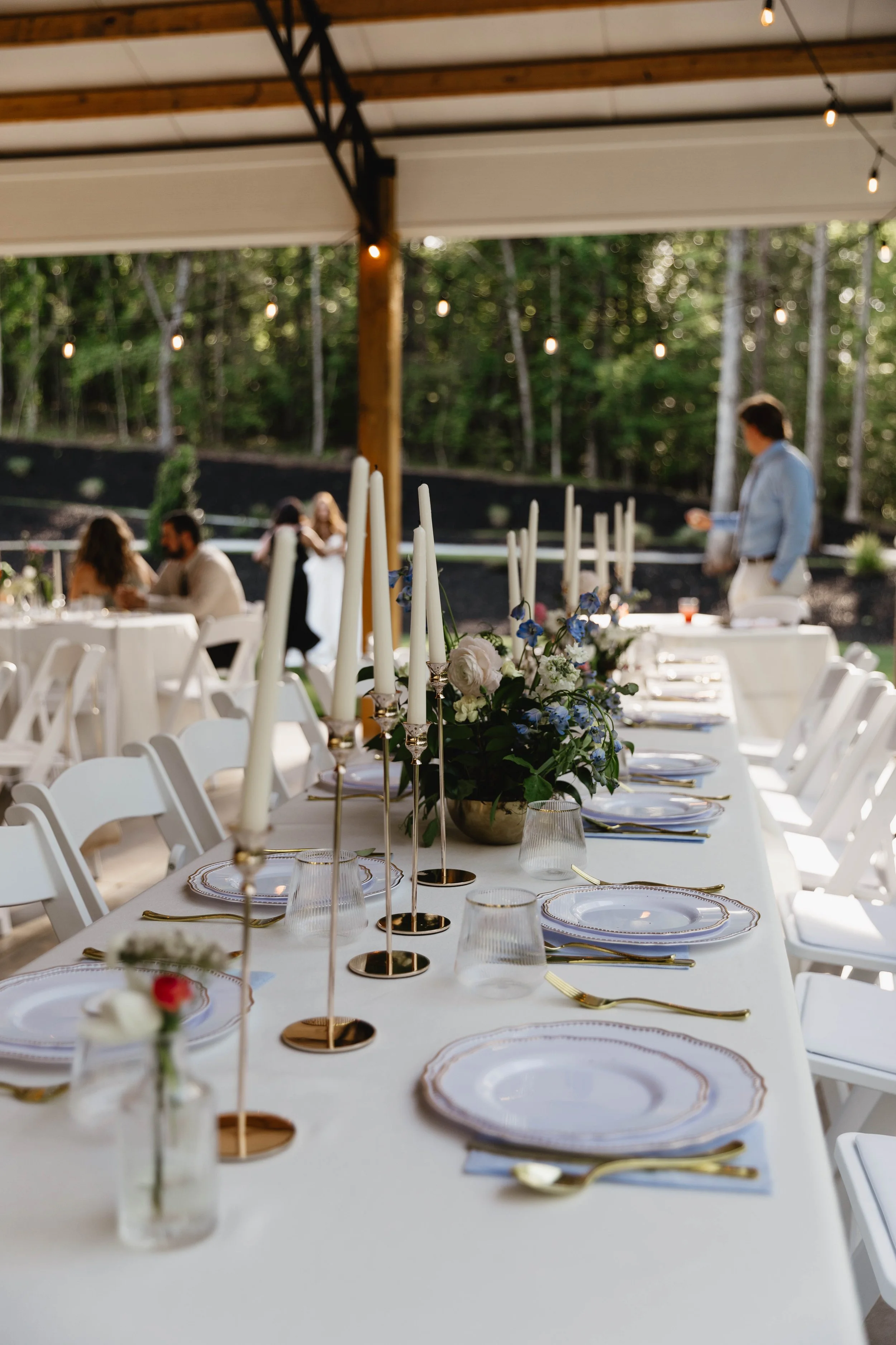 A decorated outdoor event space with a long table set for a formal dinner, featuring white chairs, floral centerpieces, tall white candles in gold holders, and gold-accented tableware. String lights are hung overhead, and there are guests and a person standing in the background.