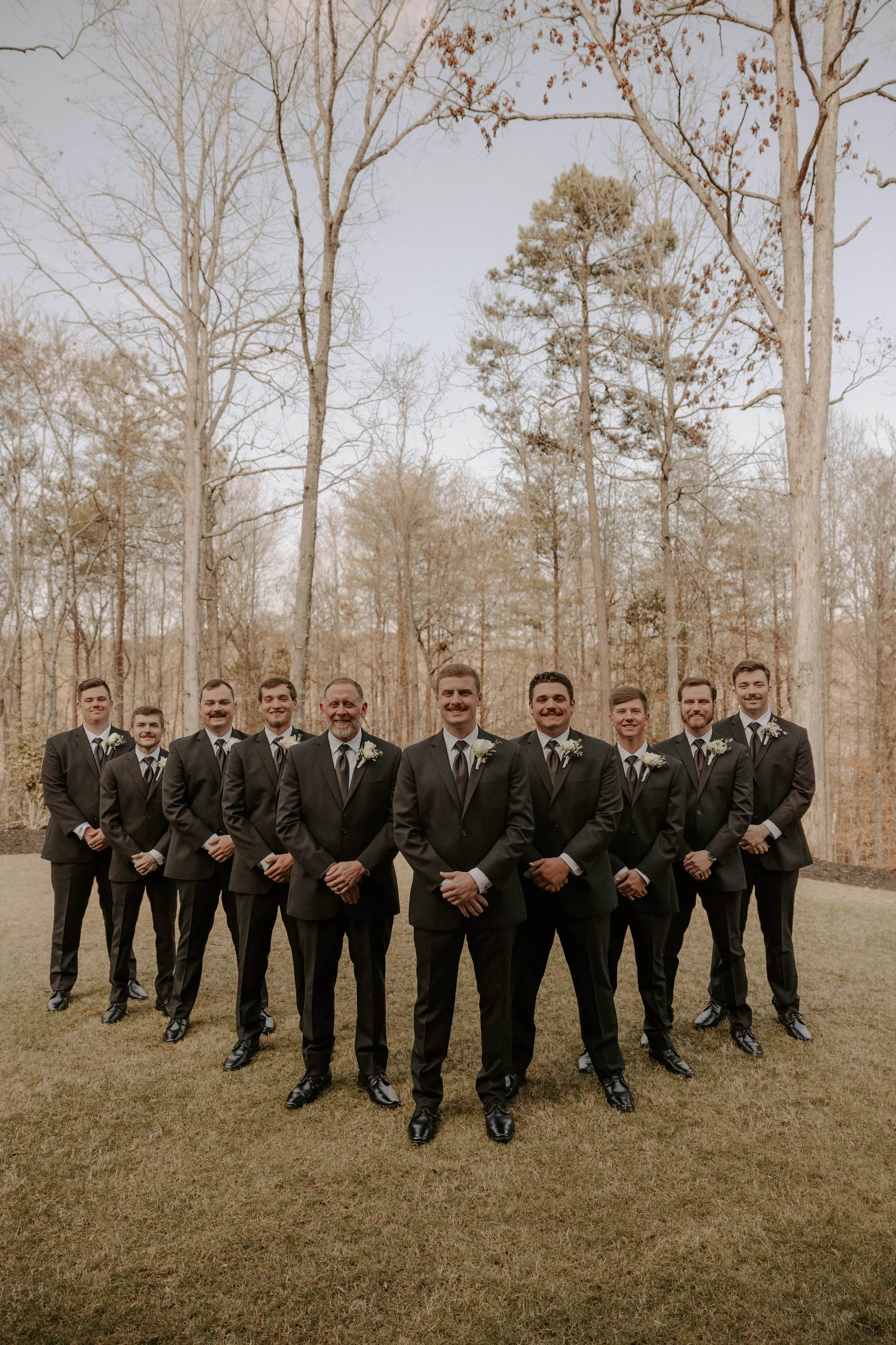 A group of men dressed in formal black tuxedos with white shirts and black ties, standing outdoors on grass with a background of leafless trees in a forested area during fall or winter.