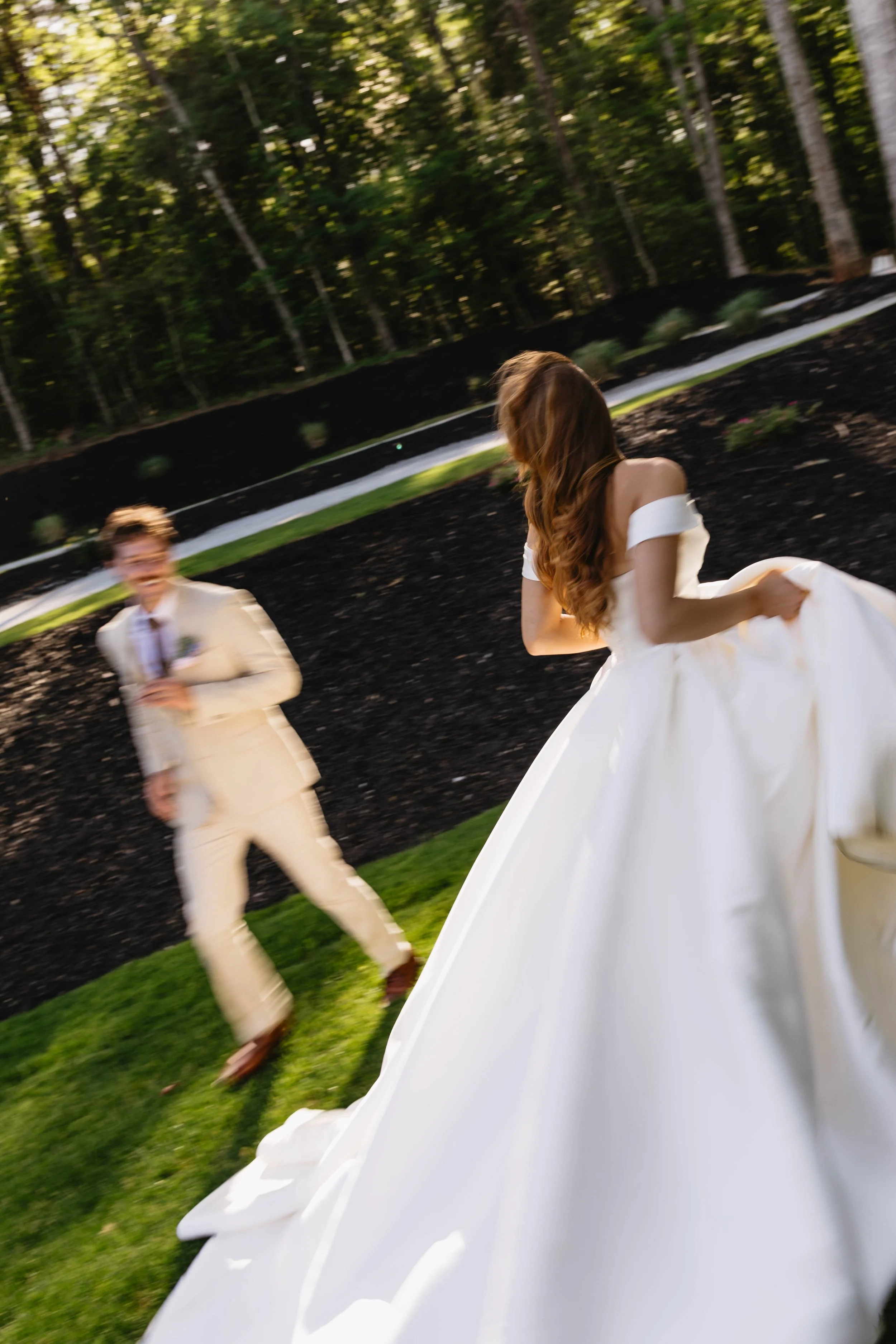 A bride in a white wedding gown and a groom in a beige suit with a white shirt and tie are outdoors on green grass, with a background of trees and black mulch. The bride's face is not visible as she looks away, while the groom is smiling and blurred in motion.