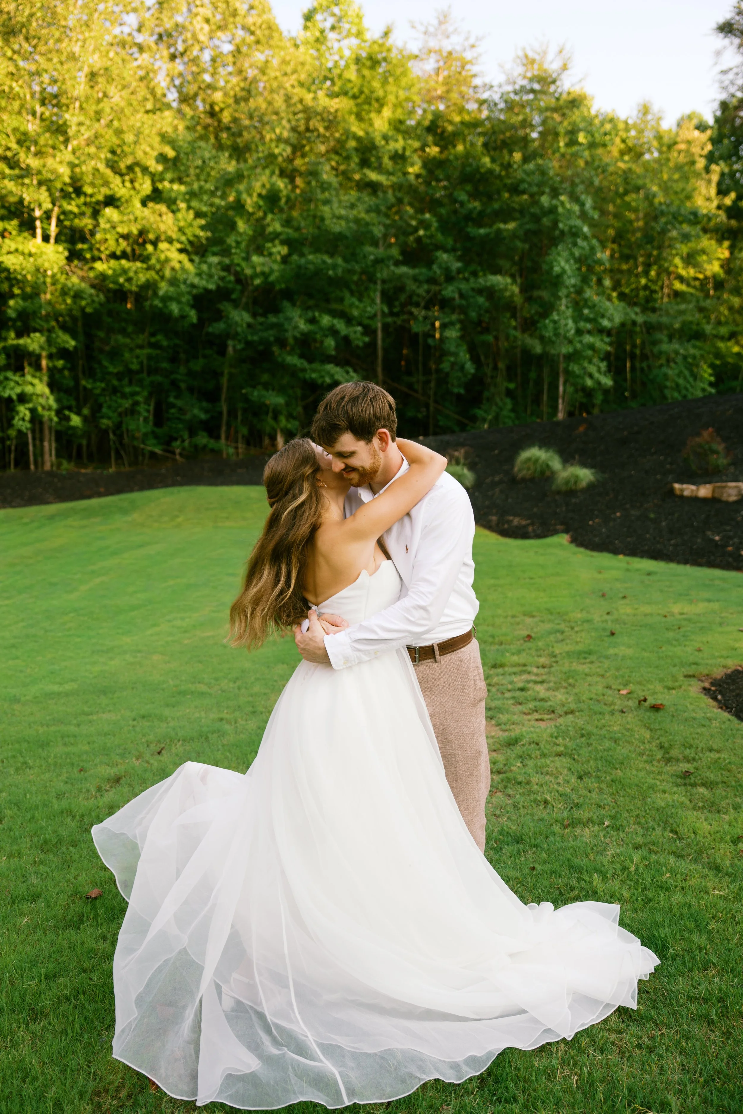 A bride and groom embrace outdoors on green grass with lush trees in the background. The bride wears a white wedding dress with a long, flowing train, and the groom wears a white shirt and brown pants. They are smiling and sharing a romantic moment.