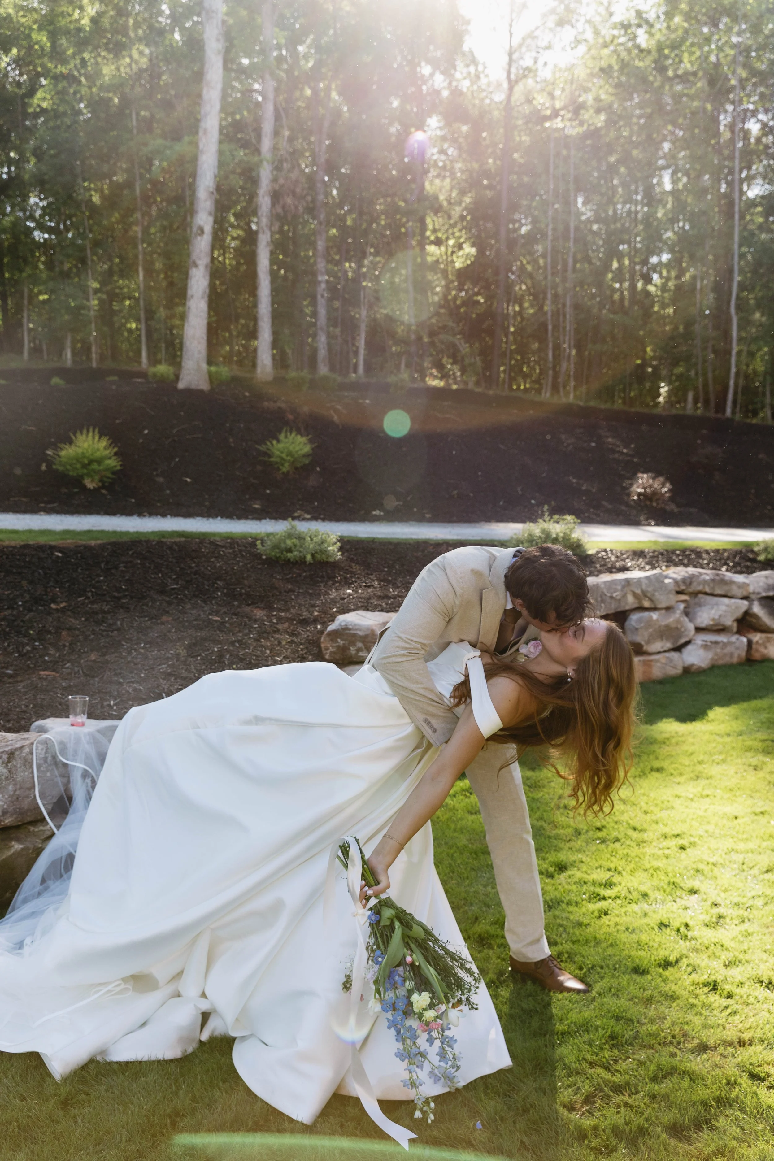 A man and a woman, dressed in wedding attire, are sharing a romantic moment outdoors during daytime. The man is leaning over the woman, who is lying on the grass with her head tilted back, holding a bouquet of flowers. The background features a lush,