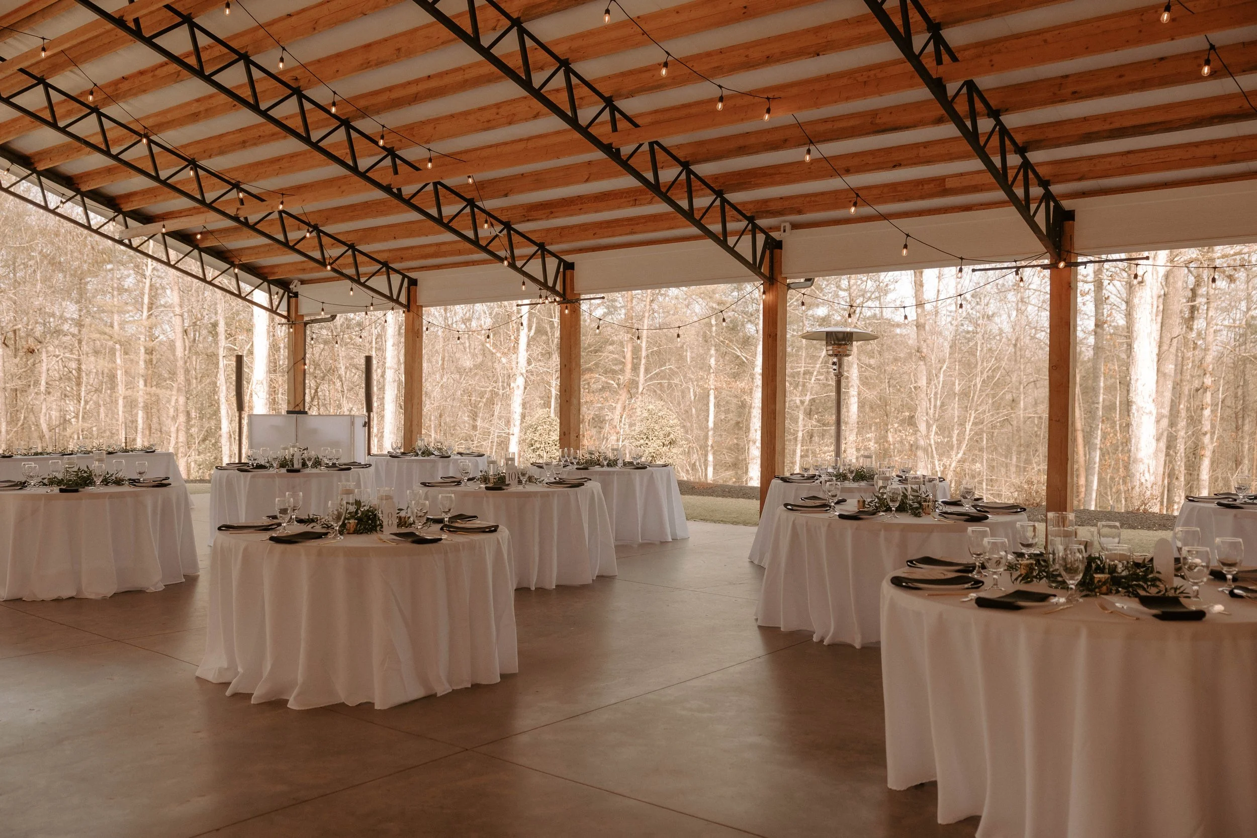An outdoor event space with round tables covered in white tablecloths, set with glassware, napkins, and small floral arrangements, under a wooden roof with string lights, surrounded by trees.