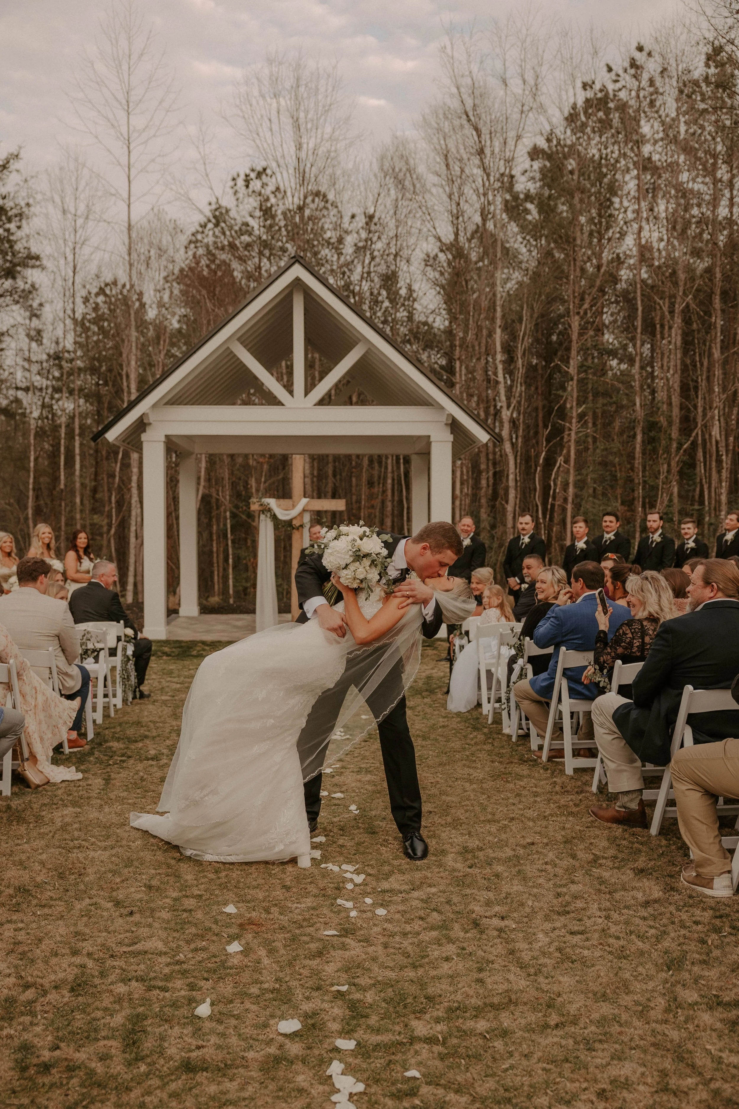 A wedding ceremony outdoors with a bride and groom dancing. The bride is wearing a white wedding gown and the groom a dark suit. Guests are seated on white chairs, and a wooden cross with a cloth is at the altar. The background contains a small white
