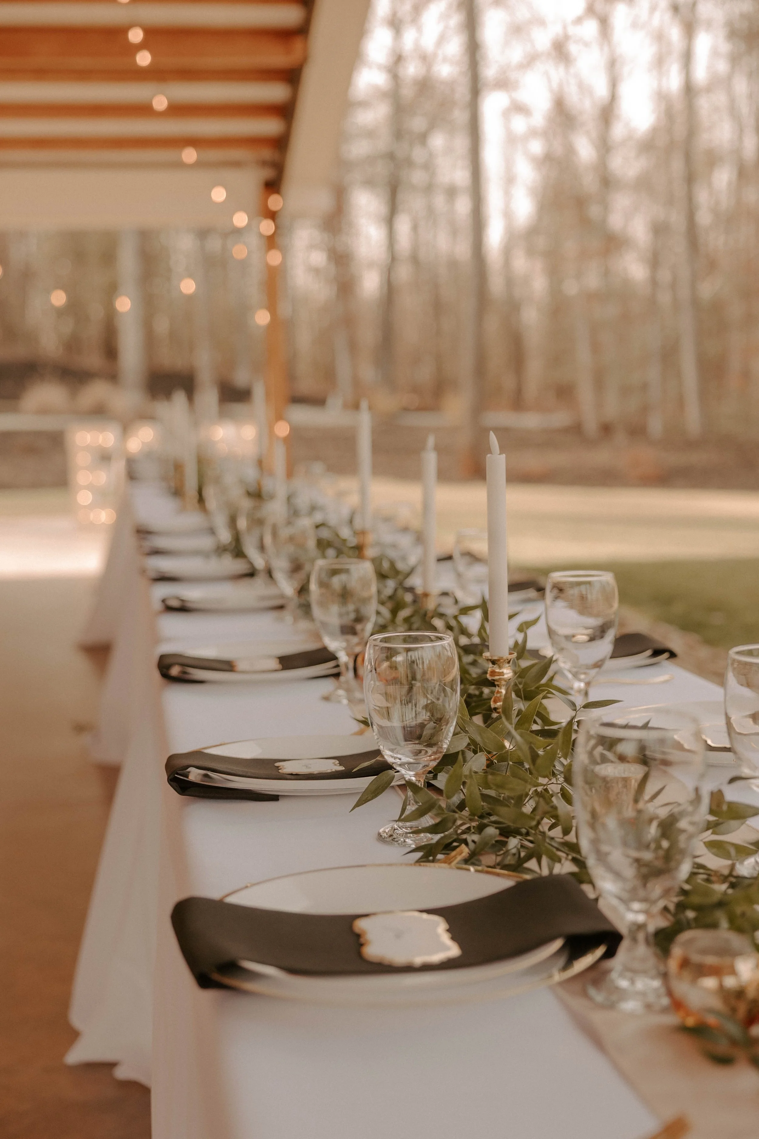 A long decorated outdoor dining table set for a celebration, with wine glasses, candles, and greenery, under string lights in a wooded area.