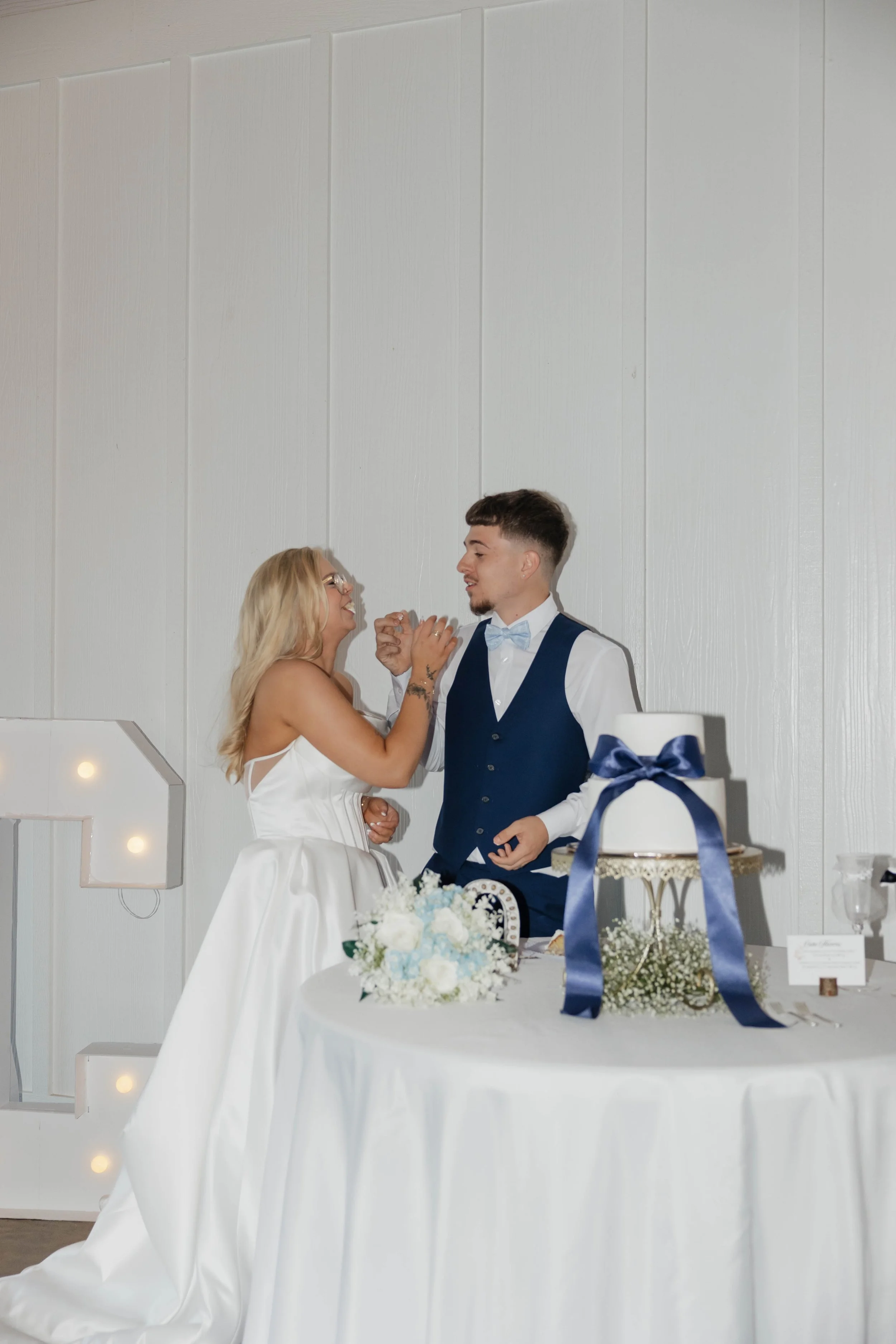 A bride and groom are at their wedding reception, holding hands and dancing together. The bride has long blonde hair and wears glasses and a white wedding dress. The groom has short dark hair and wears a blue vest, white shirt, and light blue bow tie