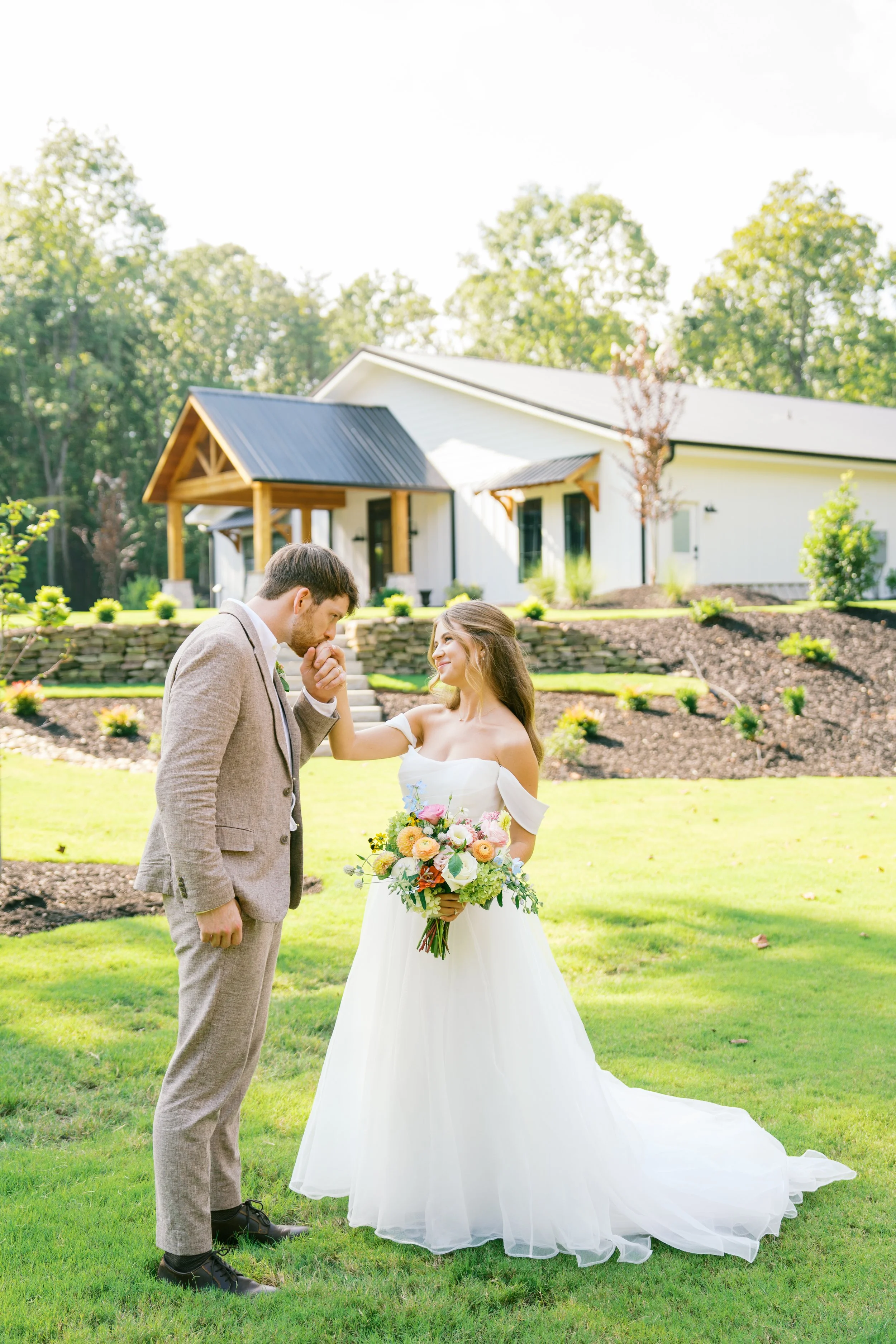 A bride and groom stand outdoors on a lawn, facing each other, with the groom holding the bride's hands. The bride is holding a large bouquet of flowers, and they are close to each other in a moment of intimacy. In the background, there is a white ho