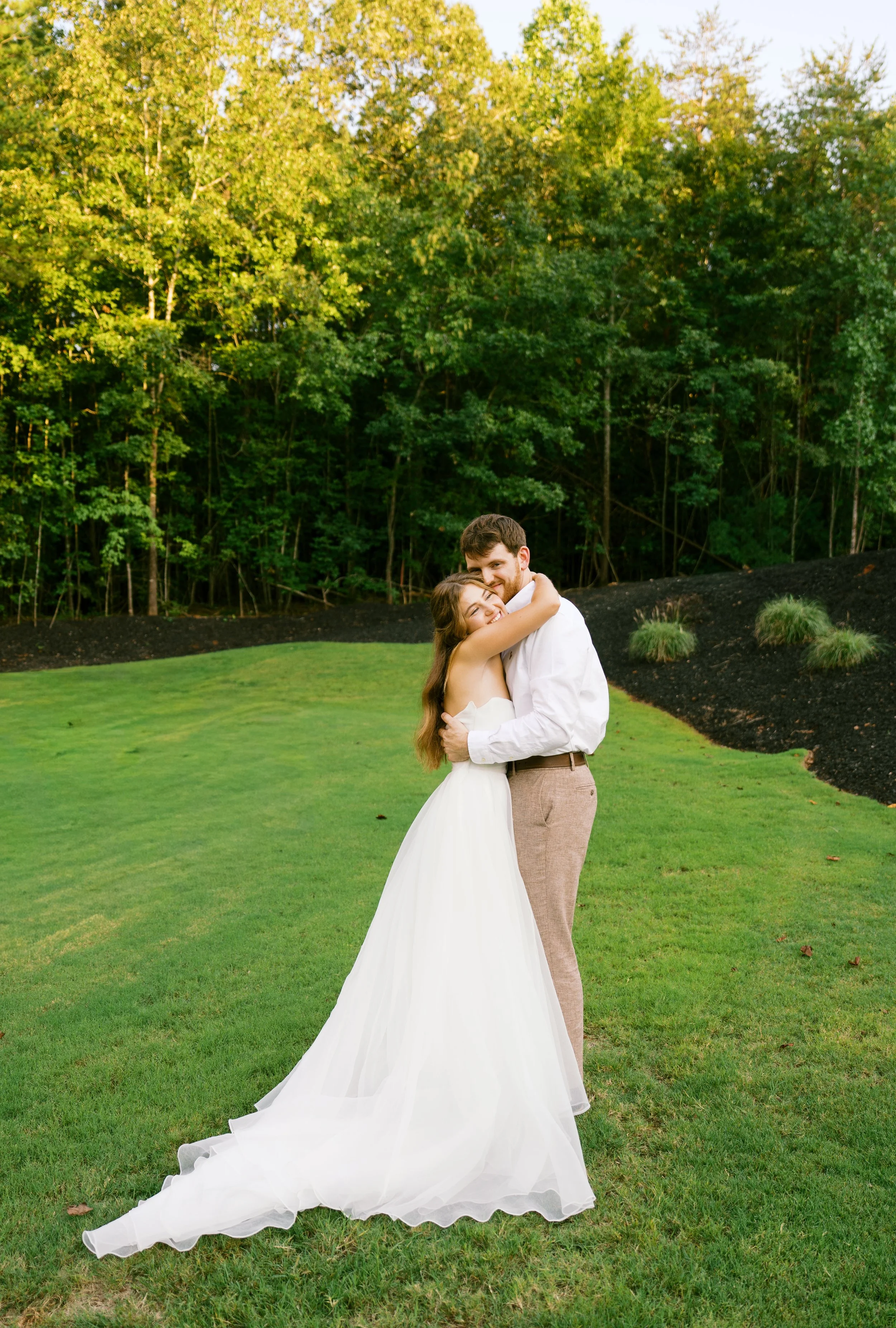 A bride and groom hugging outdoors on a green lawn with trees in the background. The bride is in a white wedding dress, and the groom is in a white shirt and beige pants.
