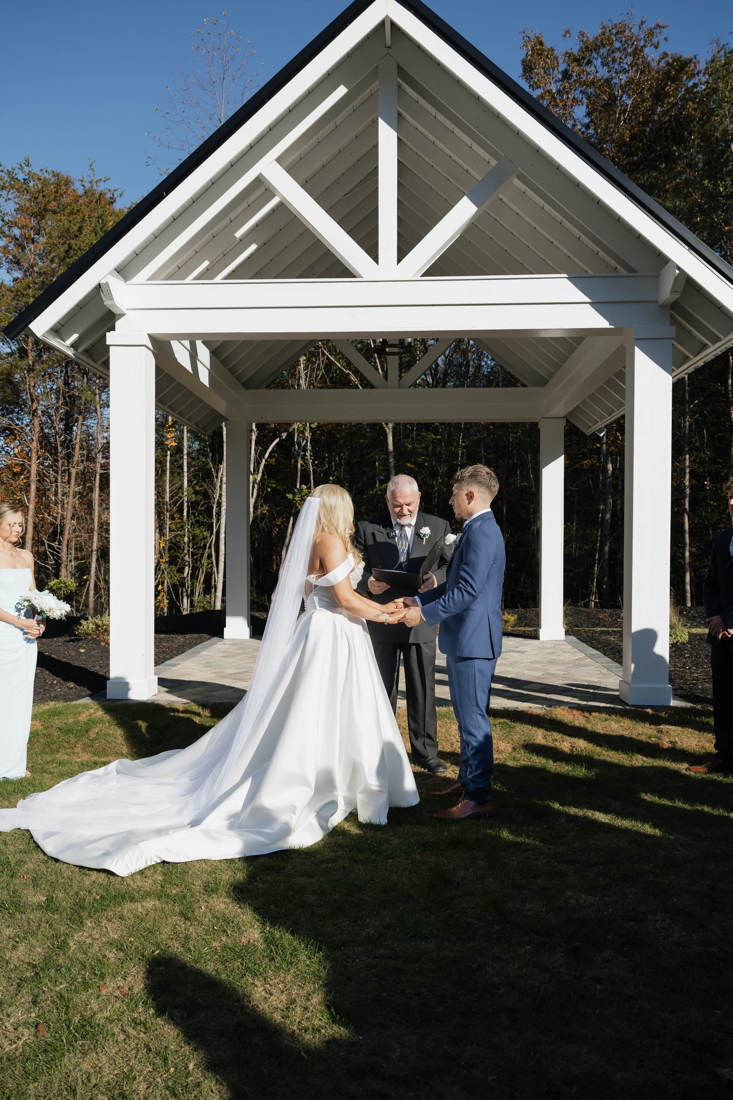 A wedding ceremony taking place outdoors under a white pavilion. A bride and groom are holding hands and facing each other, with an officiant standing between them. The bride is in a white wedding gown with a long train, and the groom is in a blue su
