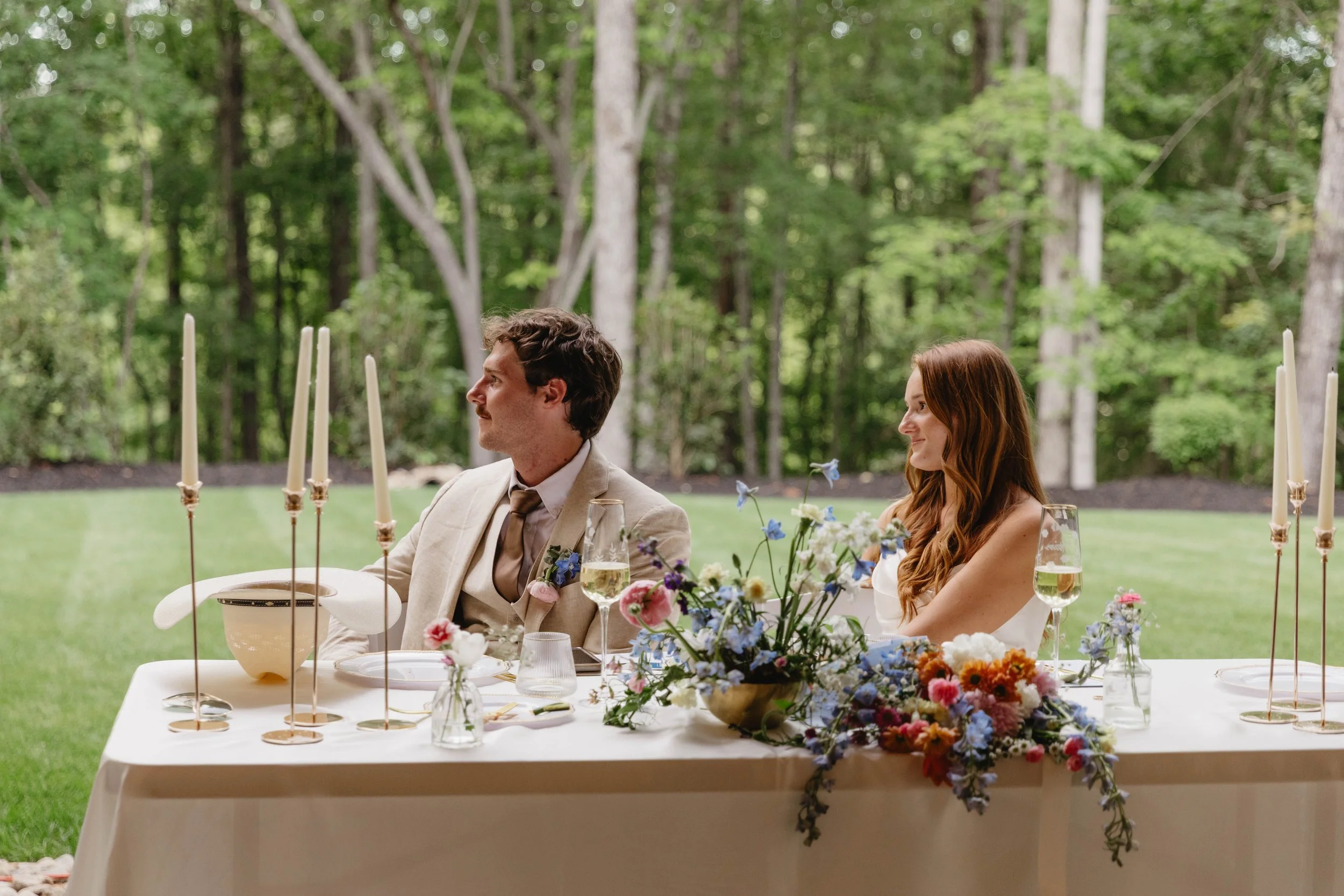 A man and woman sitting at a decorated outdoor wedding reception table with flowers and candles, surrounded by greenery.