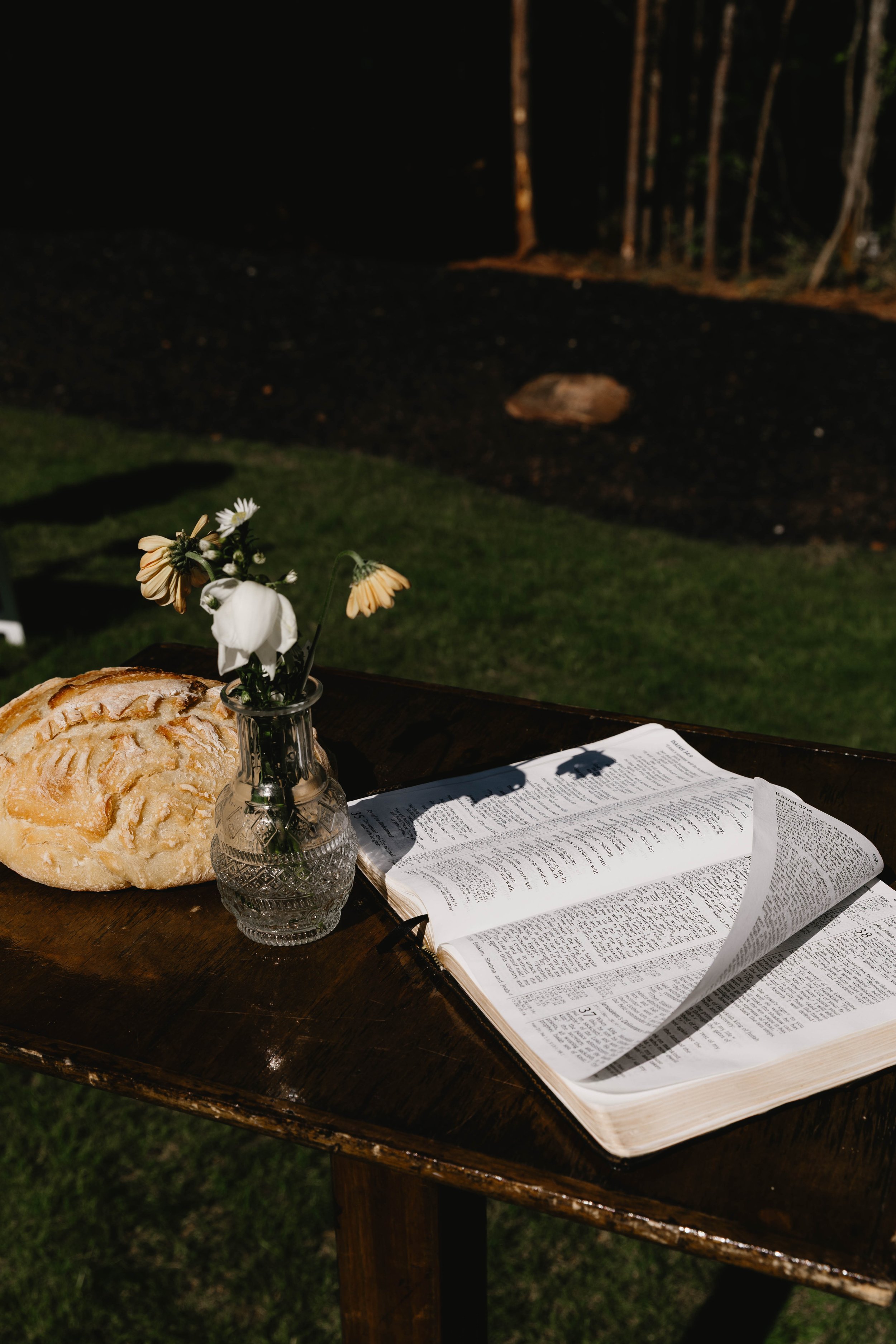 A wooden table outside with an open Bible, a loaf of bread, a small glass vase with white and yellow flowers, and a piece of paper, against a dark background of trees and grass.