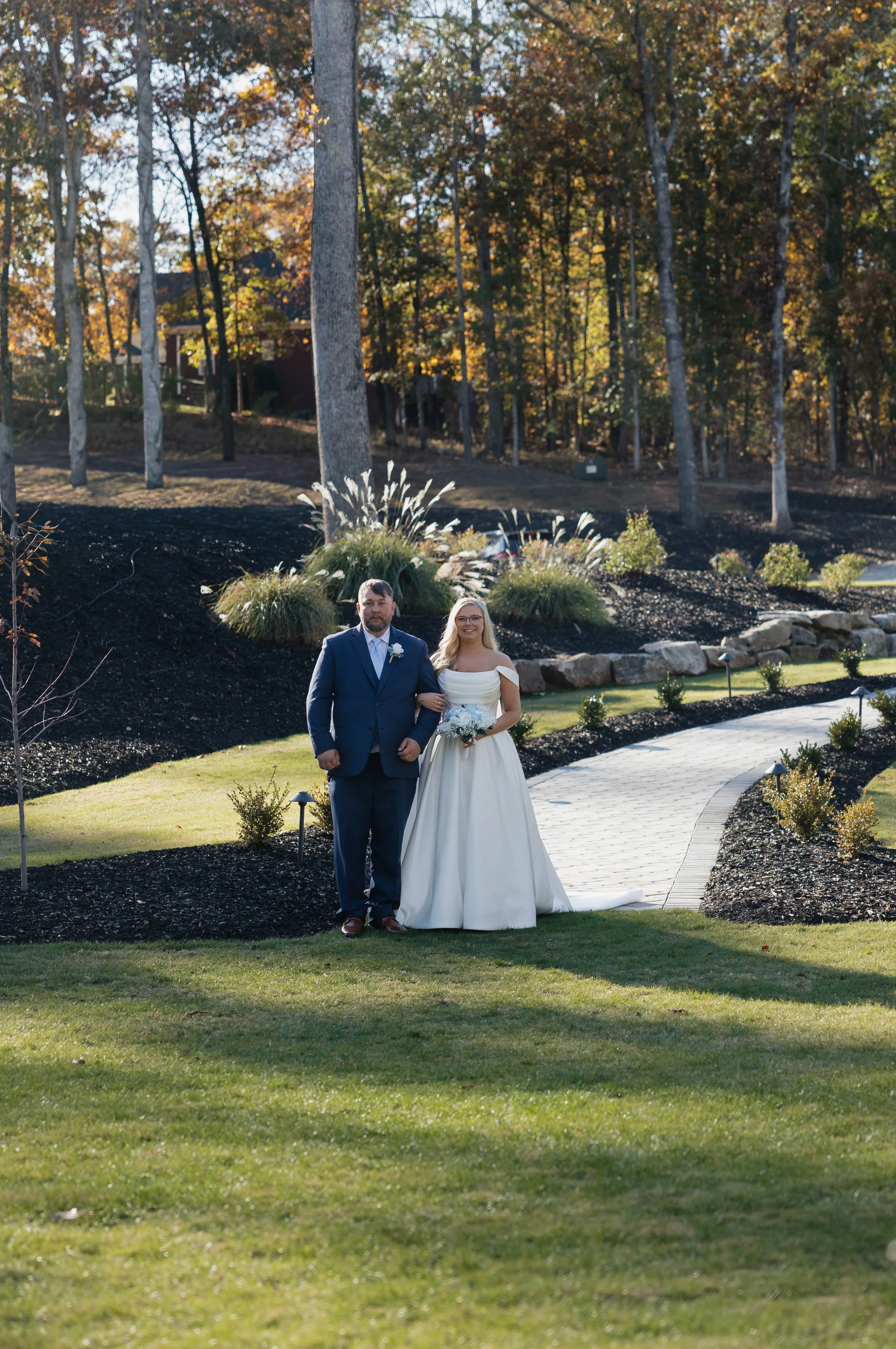 A bride and groom standing arm-in-arm outdoors on a pathway with a landscaped garden and trees in the background. The bride is wearing a white off-the-shoulder wedding gown and holding a bouquet, while the groom is in a navy suit with a white shirt a