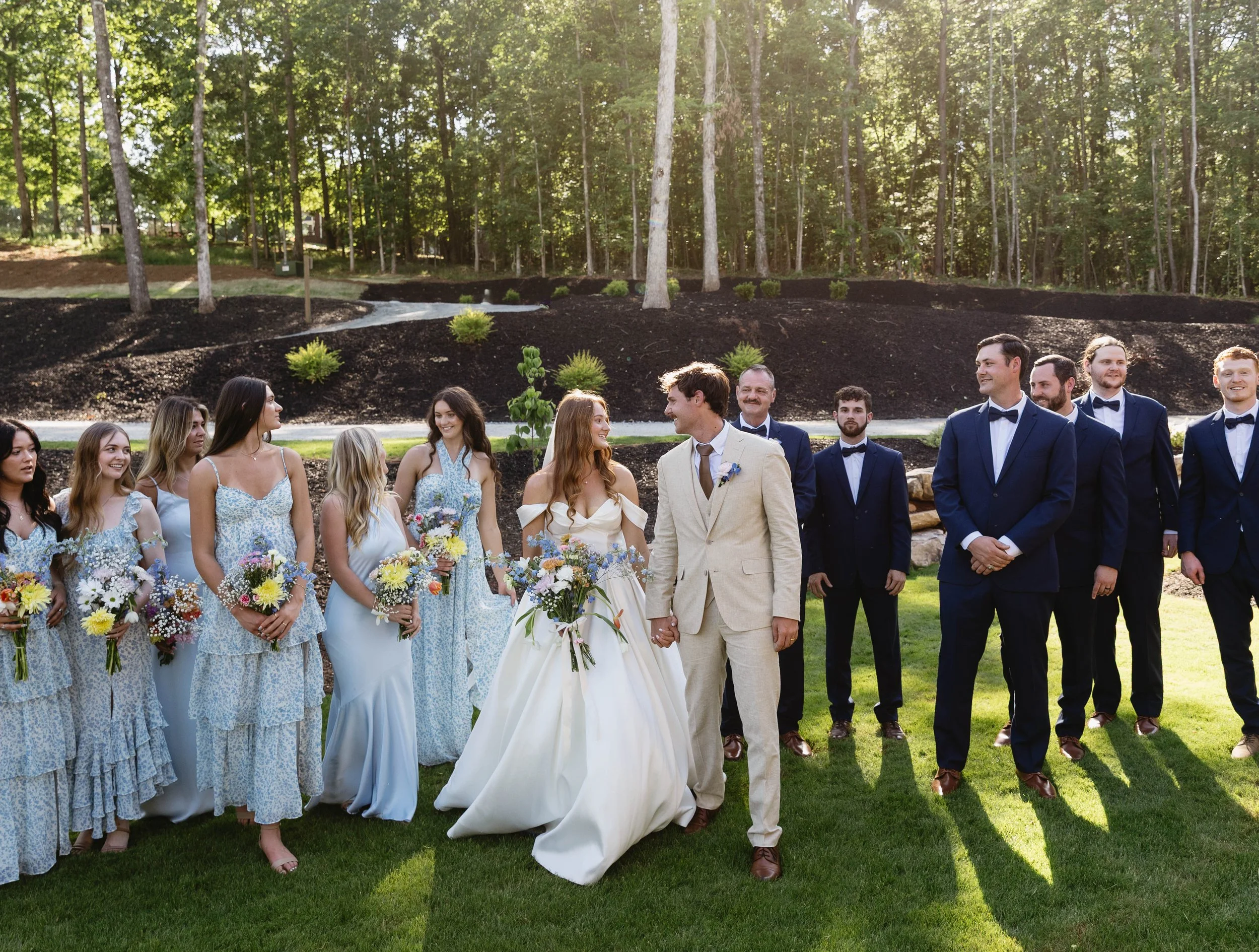 Wedding ceremony outdoors with the bride, groom, and wedding party standing on grass, surrounded by trees.