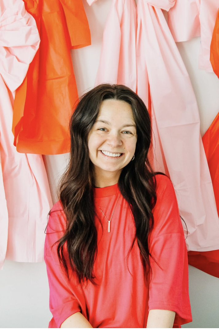 Smiling young woman with long dark hair in a red shirt, sitting in front of a colorful backdrop of orange and pink fabrics.