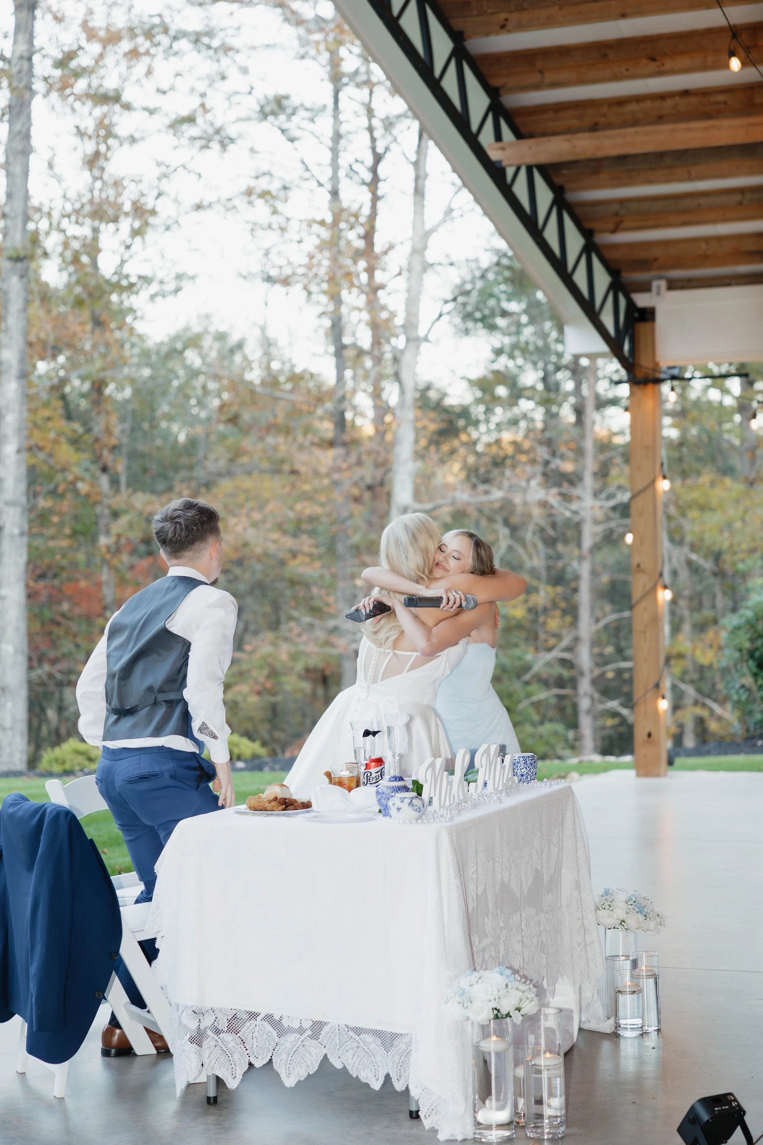 A wedding reception scene outdoors with a woman in a wedding dress hugging a blonde woman, likely the bride, at a decorated table with food and drinks, surrounded by trees with autumn foliage.
