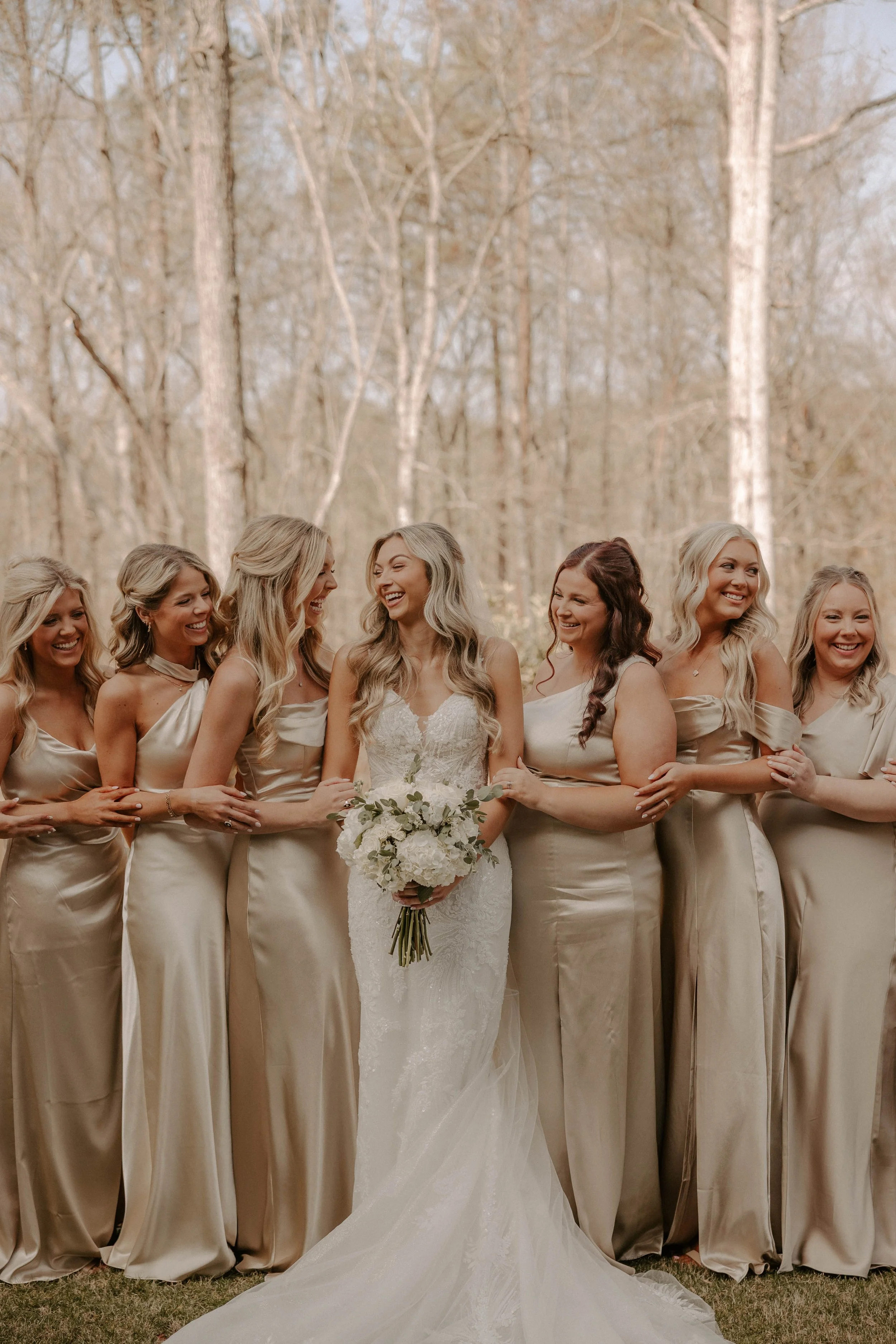 Group of women in elegant dresses, with the bride holding a bouquet, standing outdoors in front of leafless trees, smiling and interacting with each other during a wedding celebration.